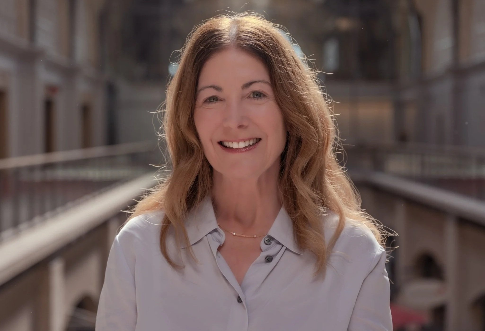 A woman with wavy, light brown hair smiling outdoors on a city street.