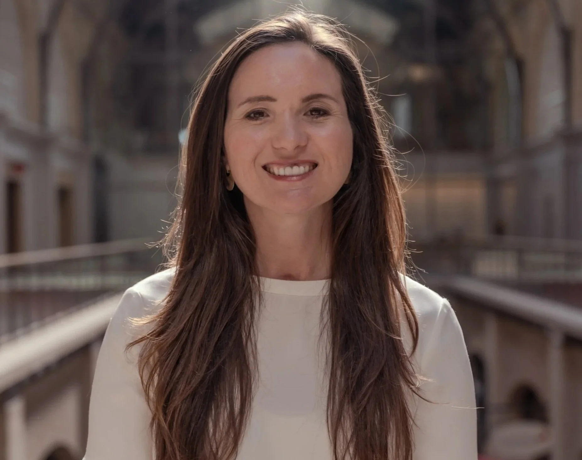 A smiling woman with long brown hair wearing a white top in an outdoor setting with a historic building in the background.
