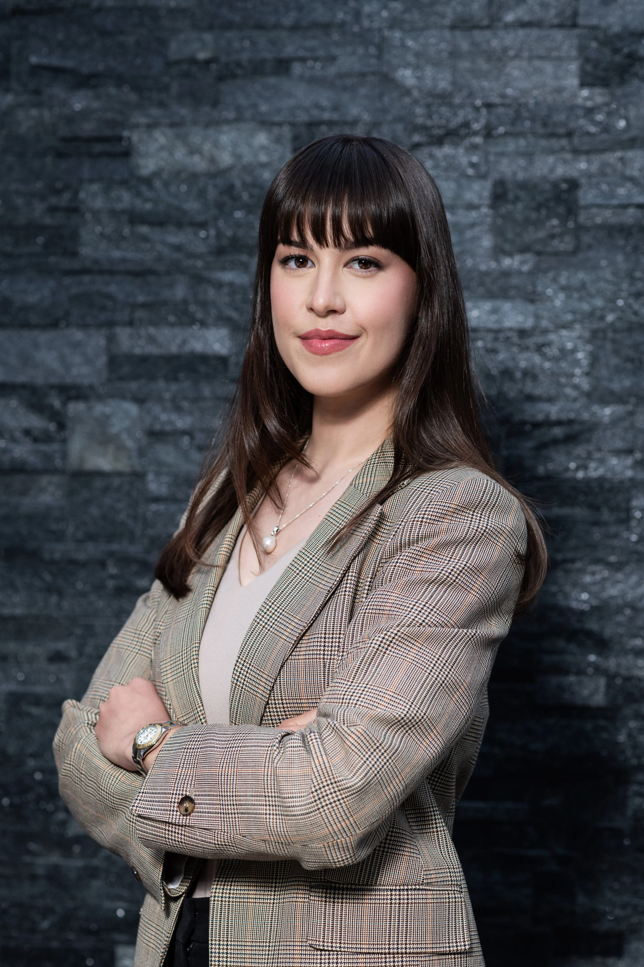A professional woman with straight dark brown hair and bangs, wearing a plaid blazer, a beige blouse, a pearl necklace, and a watch, standing with her arms crossed against a black textured wall.