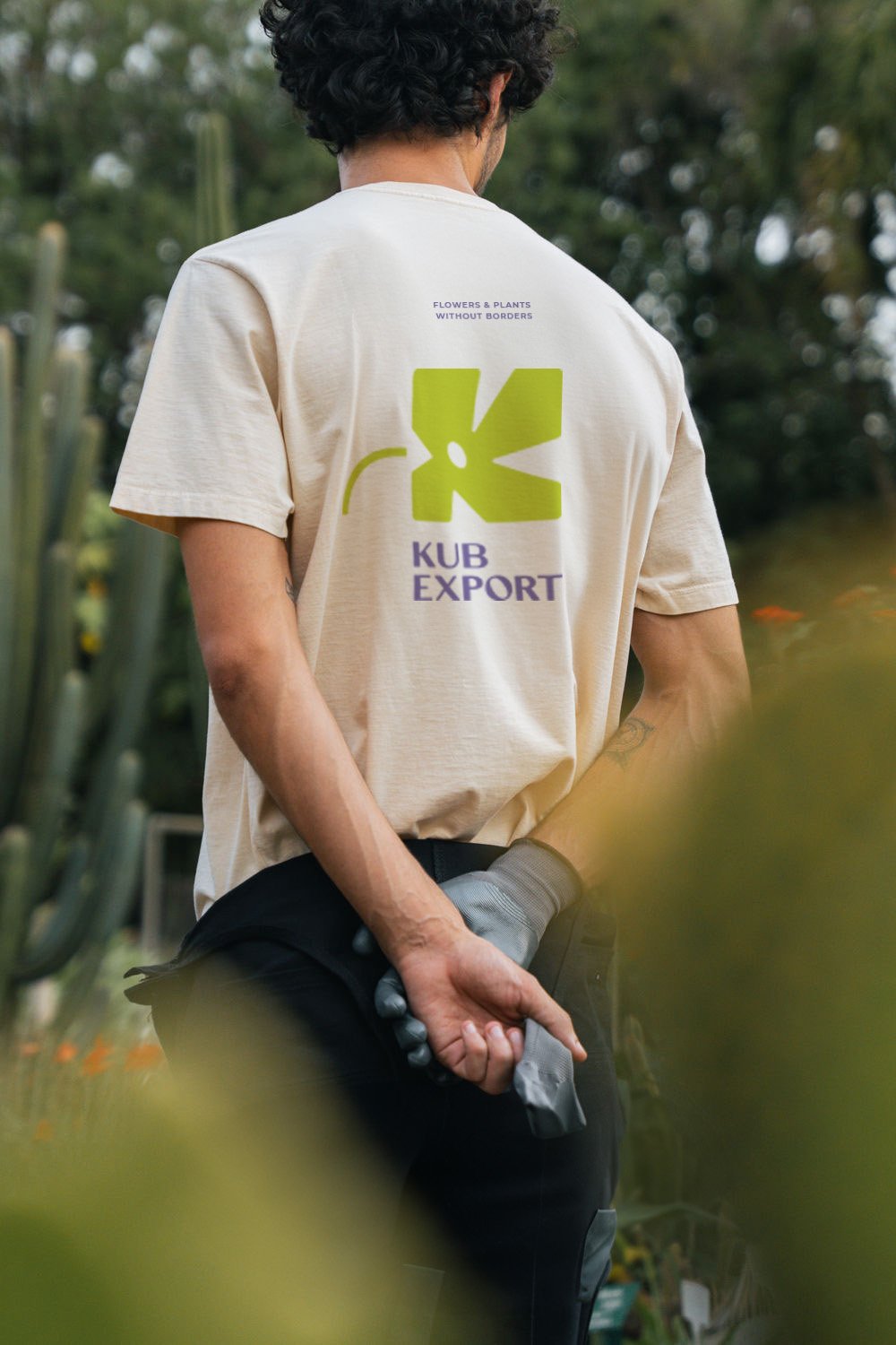 Person with curly hair wearing a beige T-shirt with a green logo and purple text on the back, standing outdoors with plants and trees in the background, holding gloves behind their back.