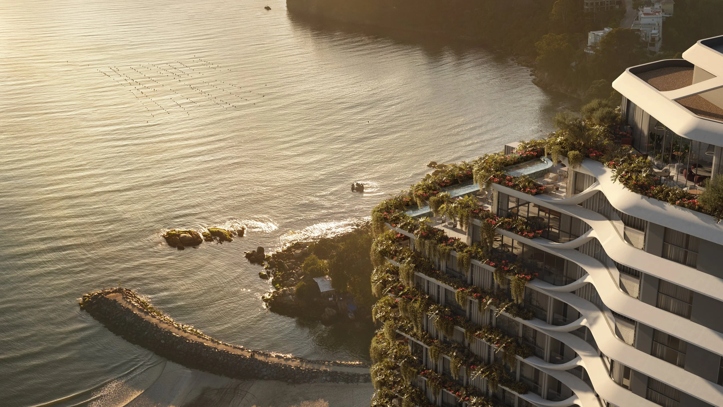 Imagem aérea de uma edifício moderno à beira-mar no litoral de Santa Catarina com varandas cheias de plantas e flores, ao lado de uma praia com água calma e um muro de pedras que delimita a orla.