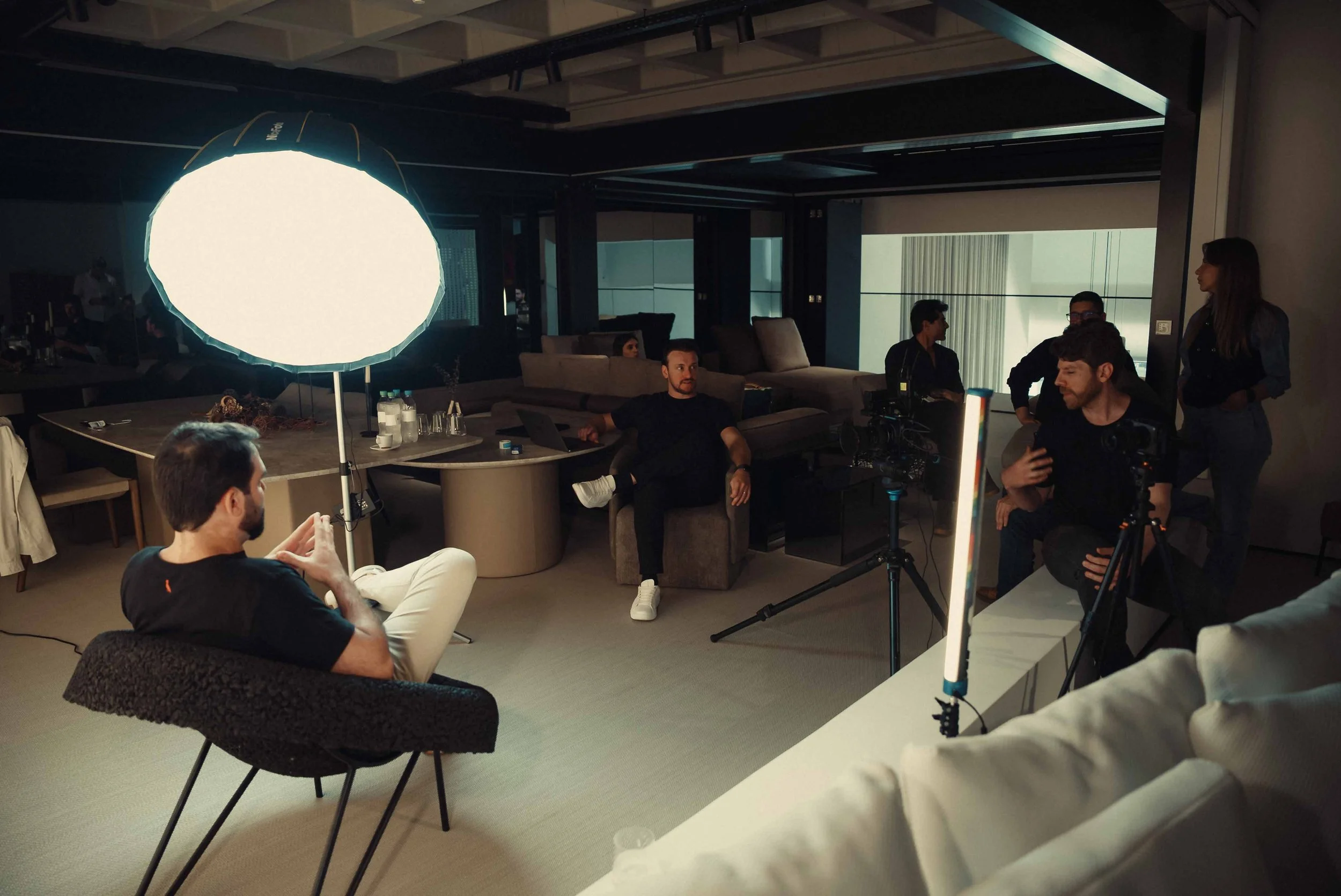 Equipe de filmagem ao redor de um set de gravação com iluminação profissional, onde uma pessoa está sendo entrevistada ou gravada.”}