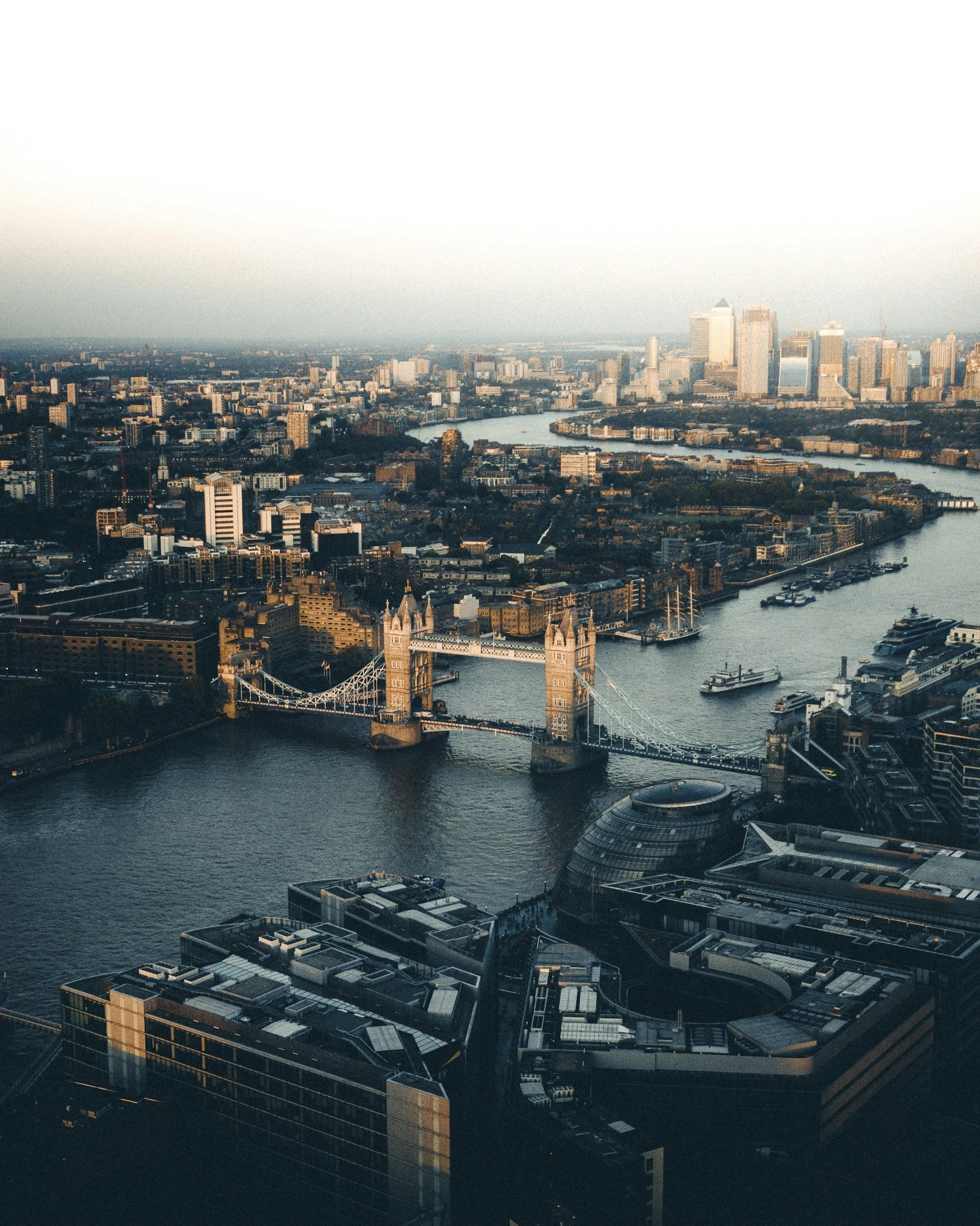 An aerial view of London featuring Tower Bridge, the River Thames, and the city skyline with various modern high-rise buildings and historic structures, taken during daytime.