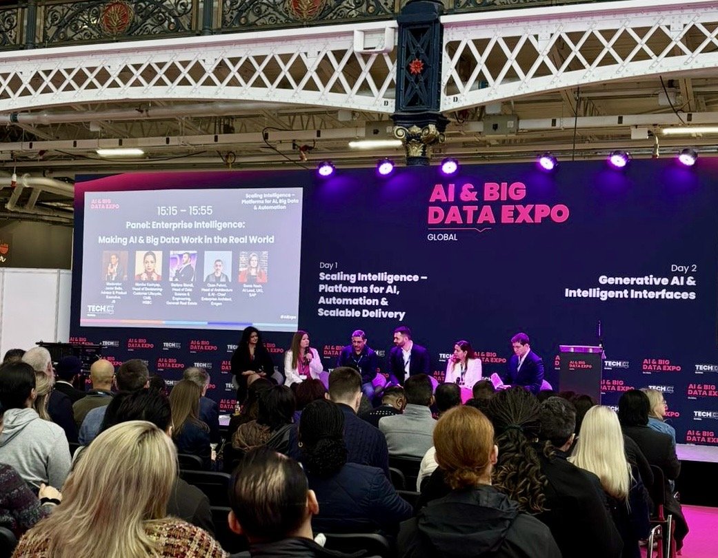 A panel discussion at the AI & Big Data Expo with six panelists on stage, a large screen behind them displaying the event theme, schedule, and topics, and an audience of attendees watching the presentation.