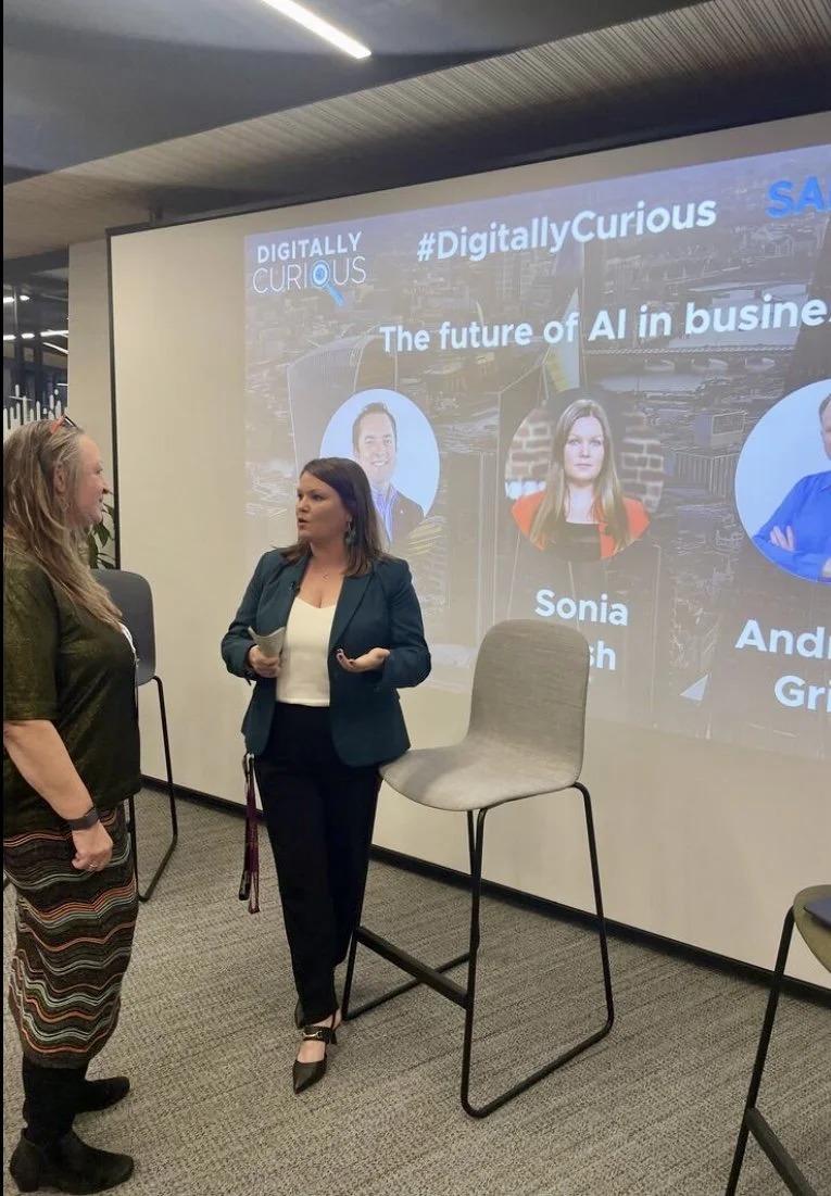 Two women engaged in conversation in front of a large presentation screen that displays the title 'The future of AI in business' along with pictures and names of speakers, inside a modern conference room.