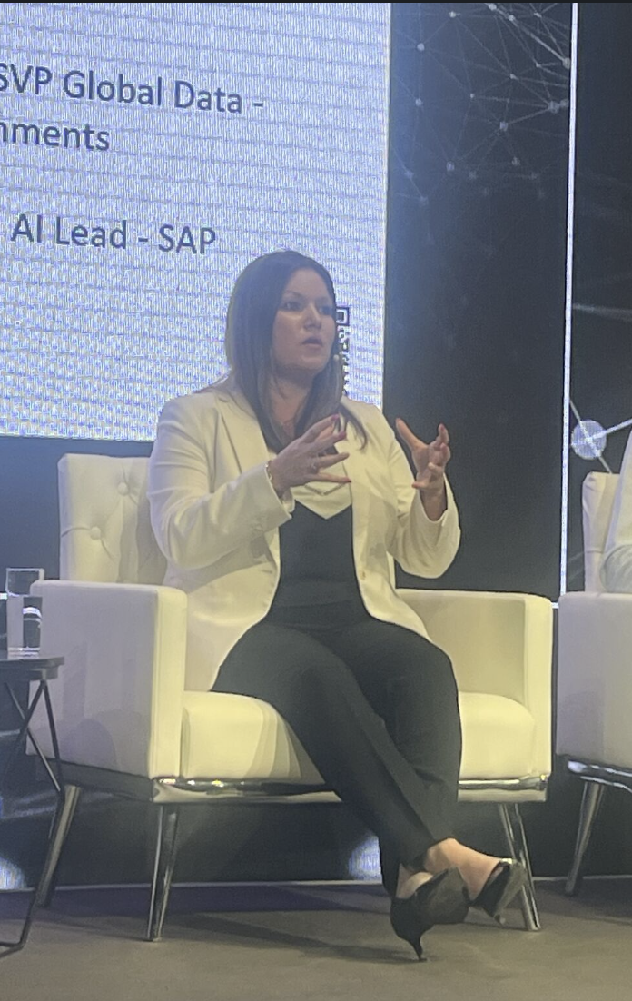 A woman in a black outfit and a light-colored blazer sitting on a white armchair on stage during a panel discussion or conference, gesturing with her hands, with a large screen displaying text and abstract graphics behind her.