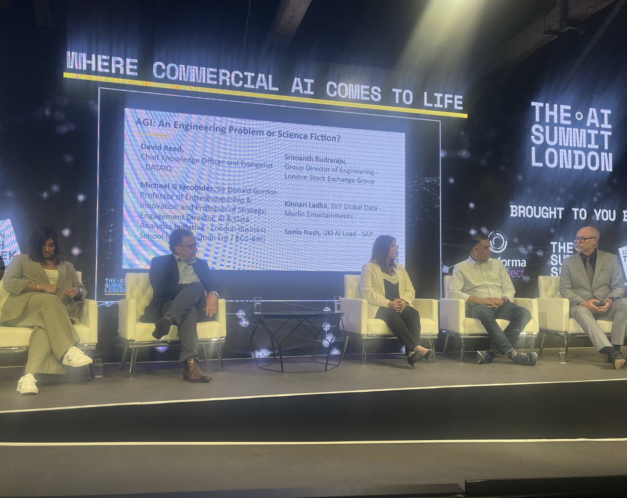 Panel discussion at The AI Summit London with five speakers seated on stage, large screen behind them displaying event details and speaker information, banner with event title and location to the right.