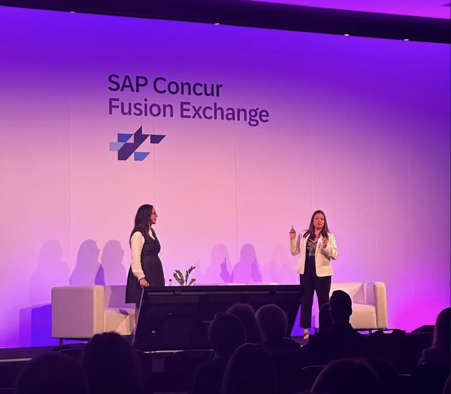 Two women on stage at SAP Concur Fusion Exchange, one speaking and the other listening, with a backdrop displaying the event name and logo, in a conference setting