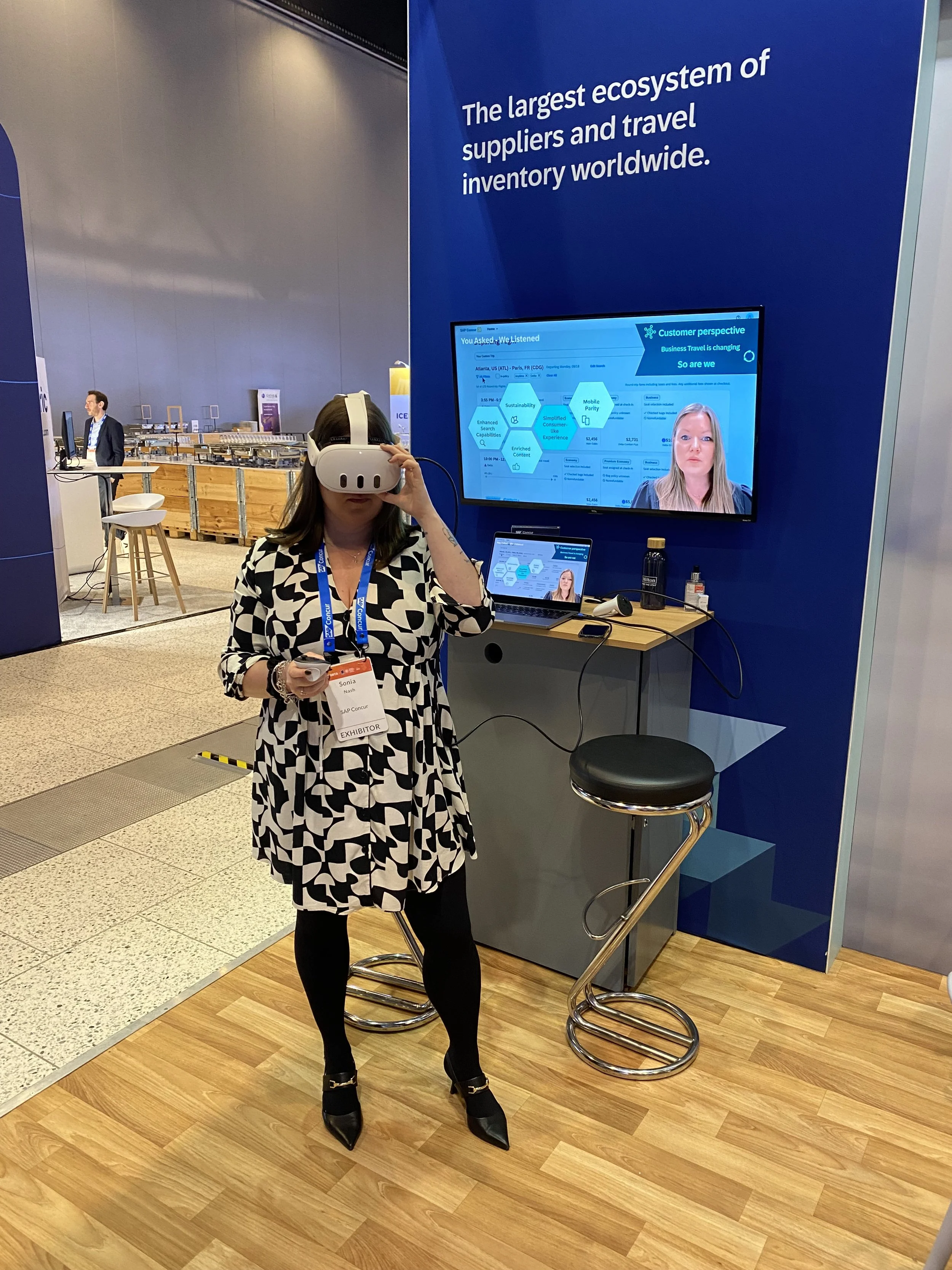 A woman with a virtual reality headset standing in front of a display at a conference, with a large screen behind her showing a presentation and a laptop on a small table.