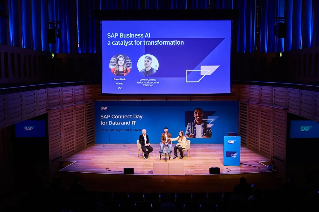 Stage at a conference with three panelists seated on chairs and a large screen behind them displaying information about SAP Business AI and SAP Connect Day.
