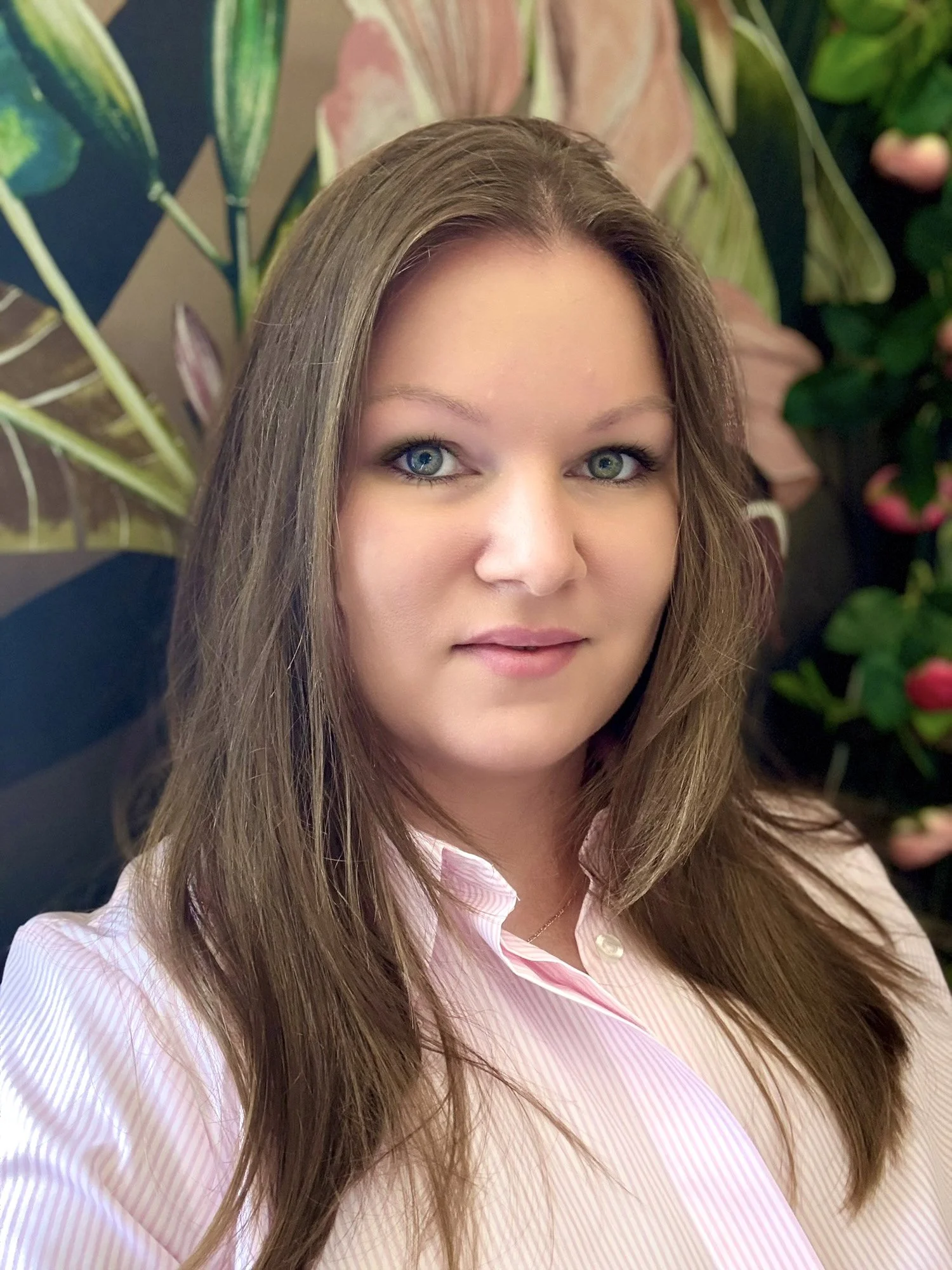 A woman with long brown hair and blue eyes wearing a pink and white striped blouse, standing in front of a background of lush green and pink plants.