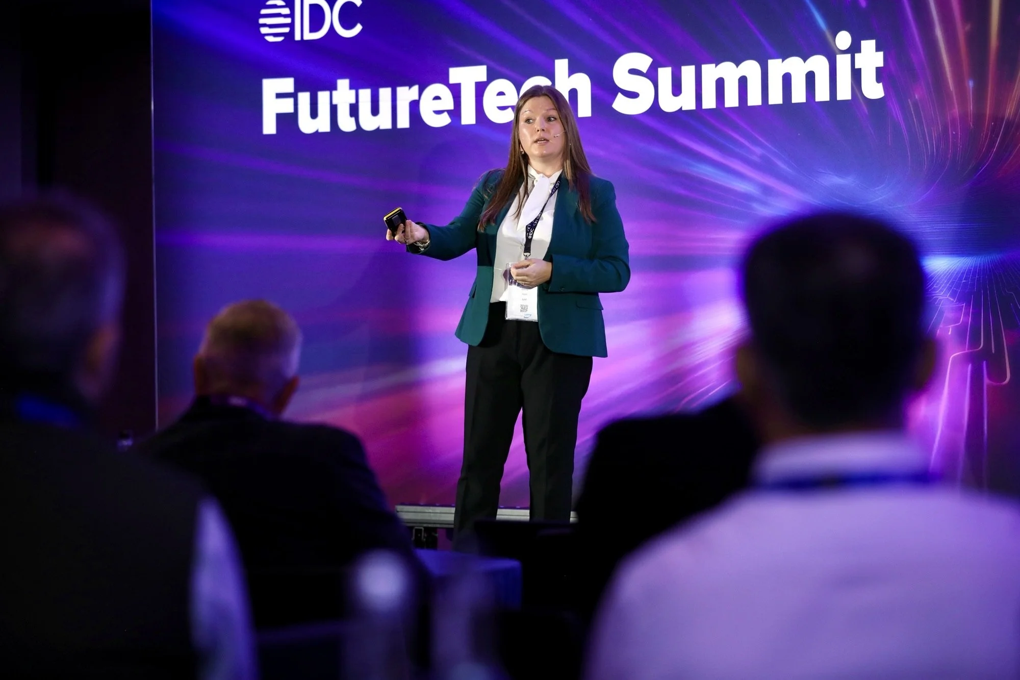 A woman giving a presentation on stage at the IDC FutureTech Summit, with a large colorful digital graphic background and seated audience members in front.