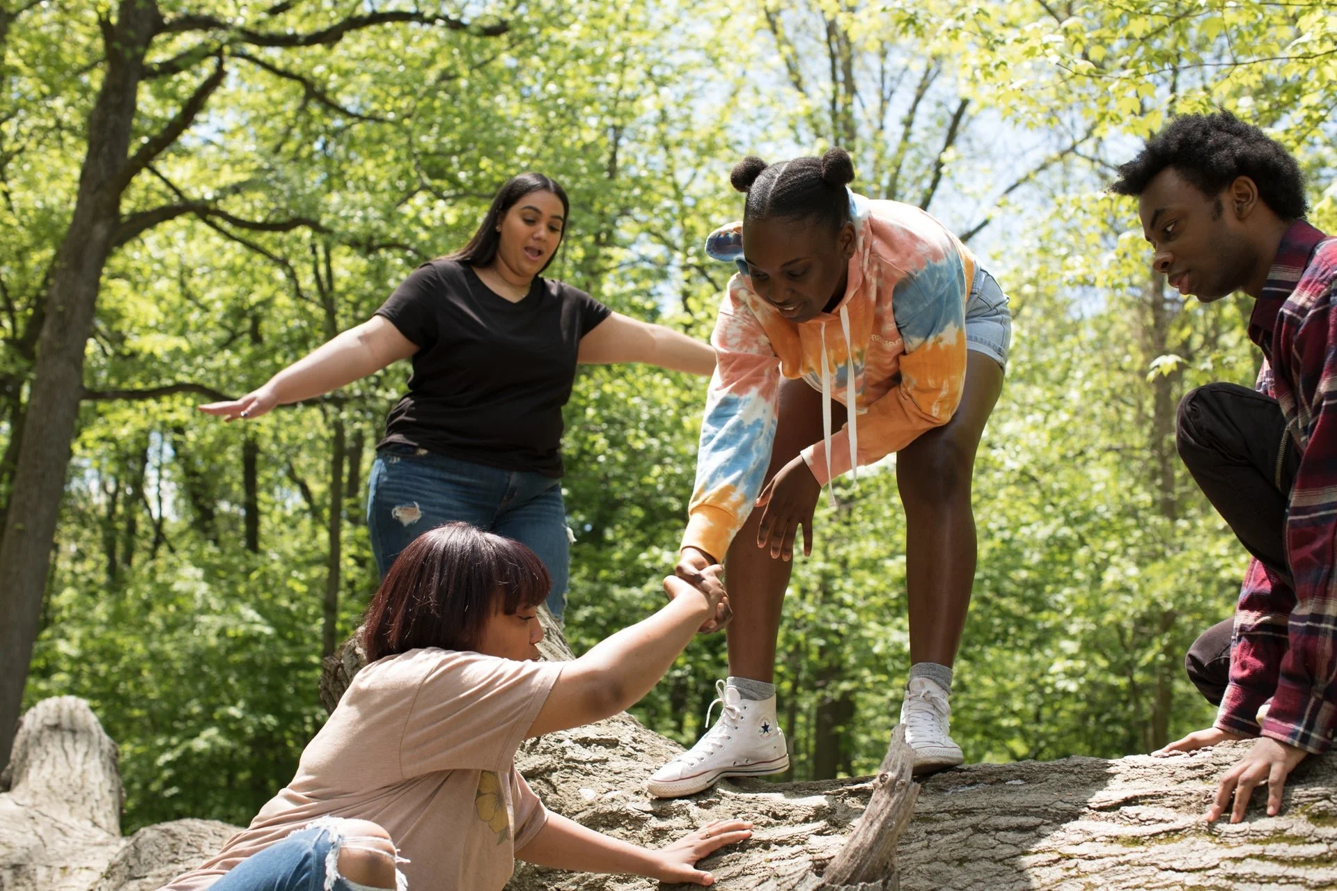 Group of five friends helping each other climb over fallen log in forest.