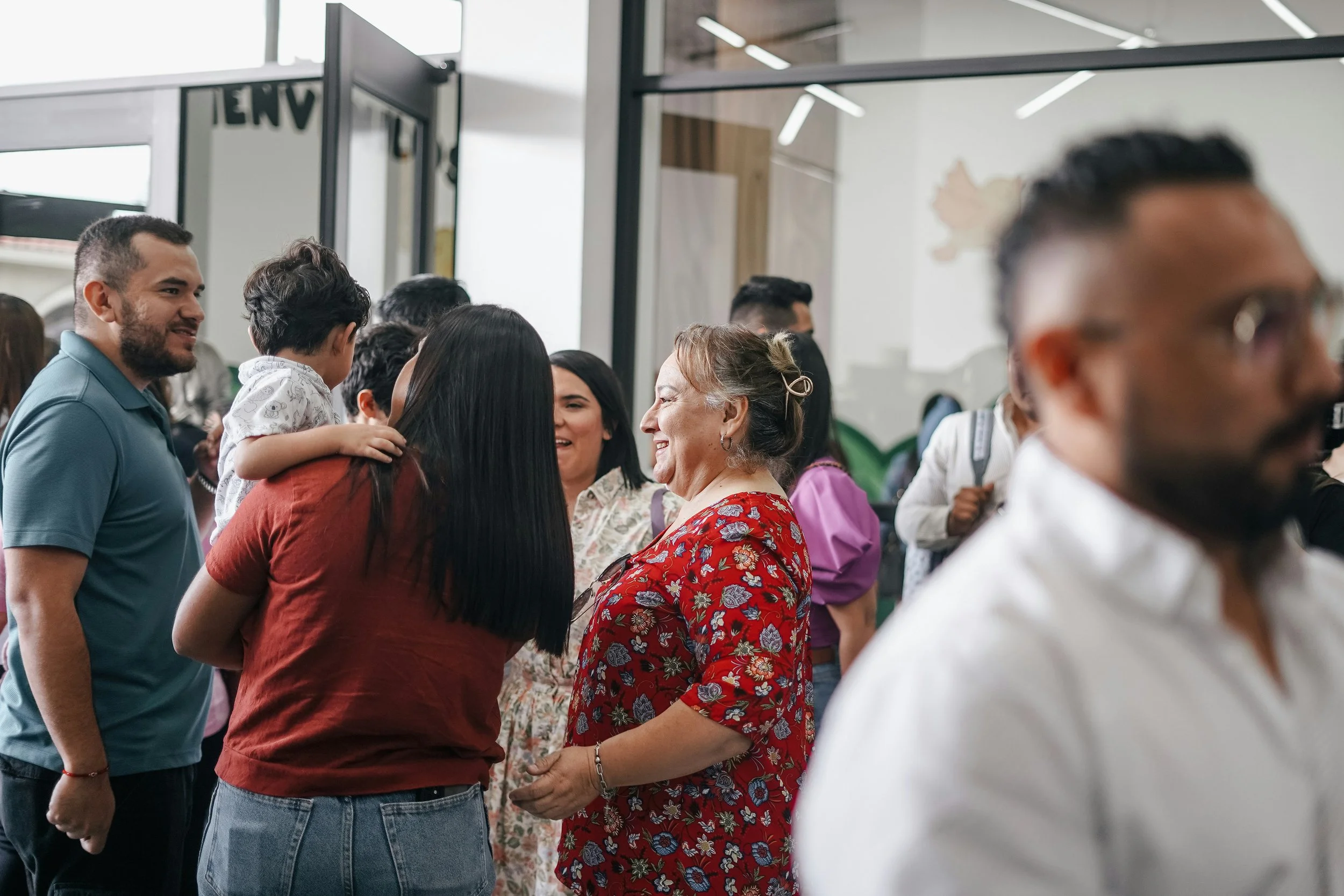 Group of people socializing indoors, smiling, and talking, including a woman in a red floral dress and a young girl on a woman's shoulder.
