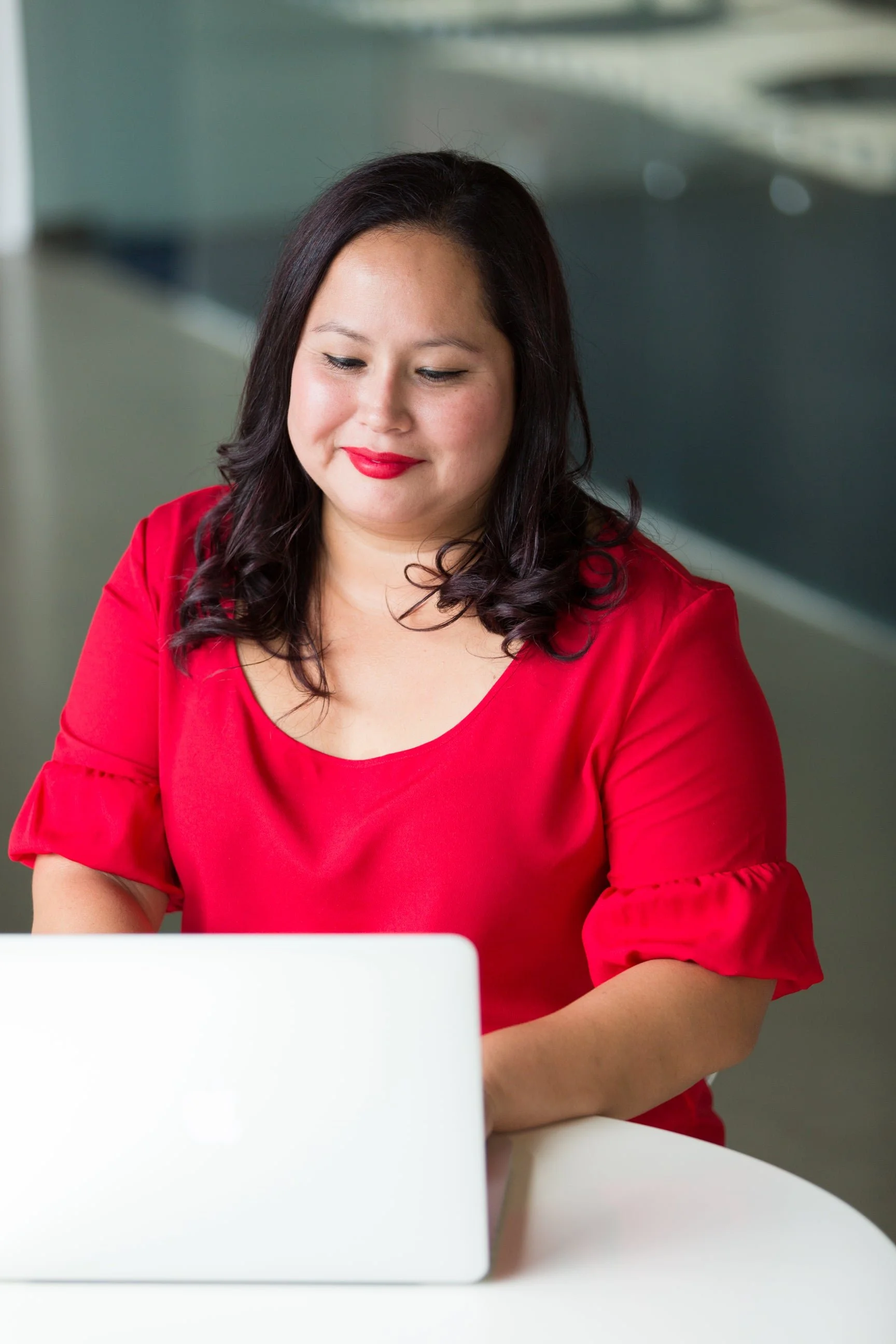 A woman with dark hair and red lipstick, wearing a red blouse, sitting at a white table and looking at a silver laptop.