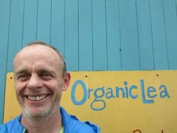 Smiling man standing in front of a yellow sign that reads "Organic Lea" against a blue wooden wall.