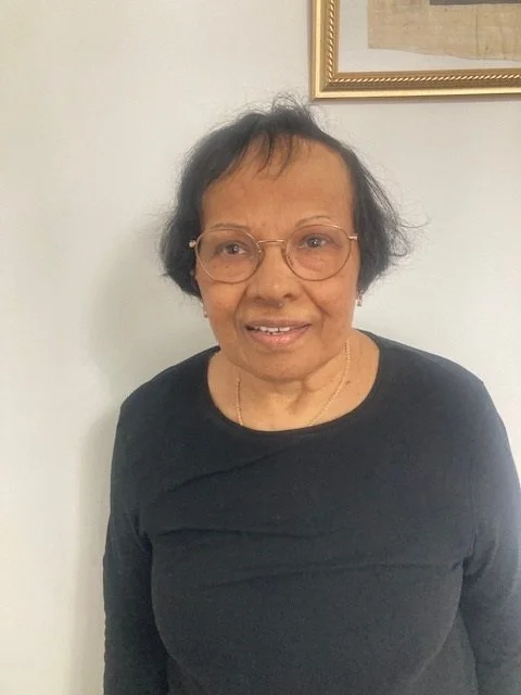 Older woman with short black hair, glasses, wearing a black top, standing indoors against a white wall with a framed picture in the background.