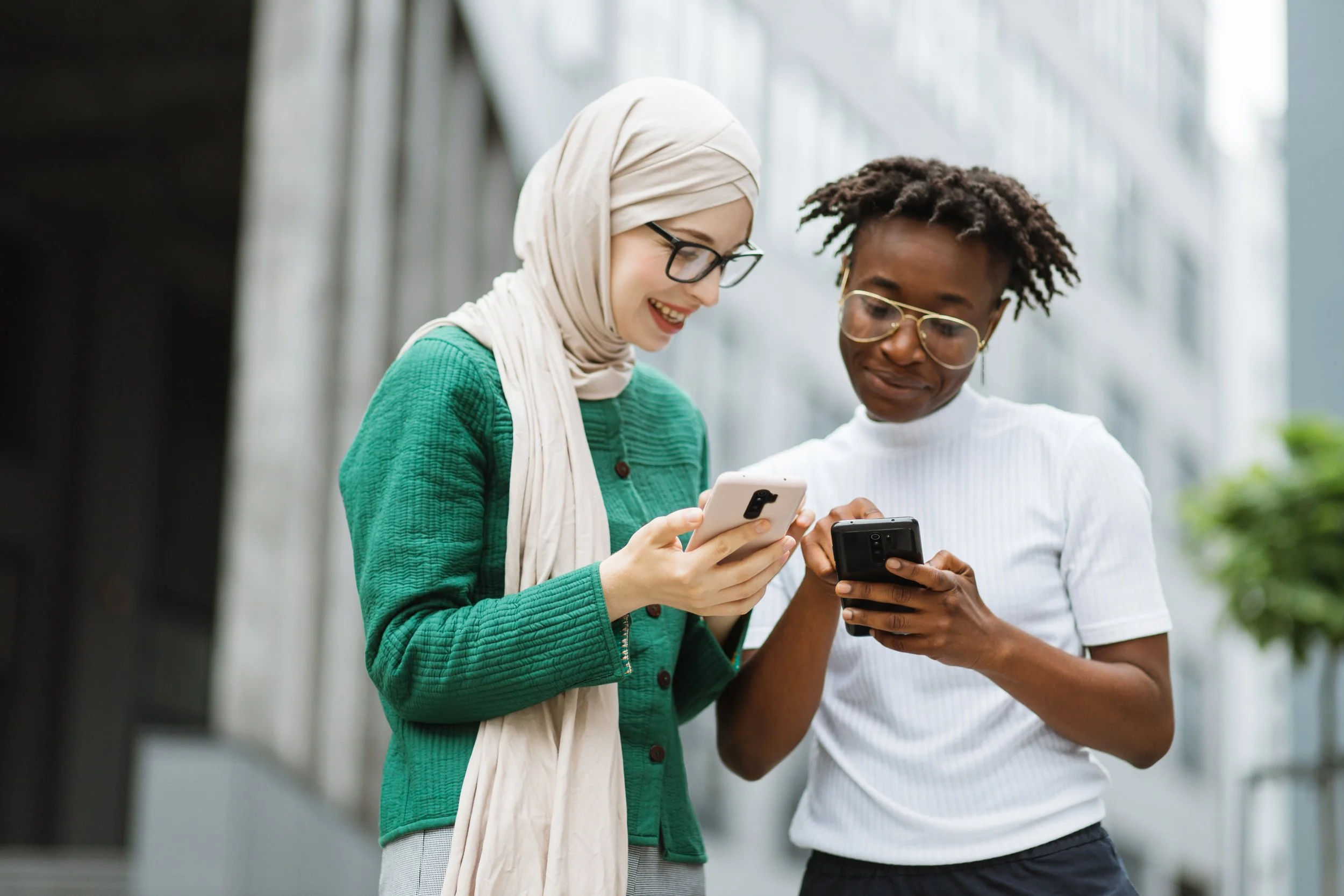 Two women standing outdoors, looking at their smartphones and smiling.