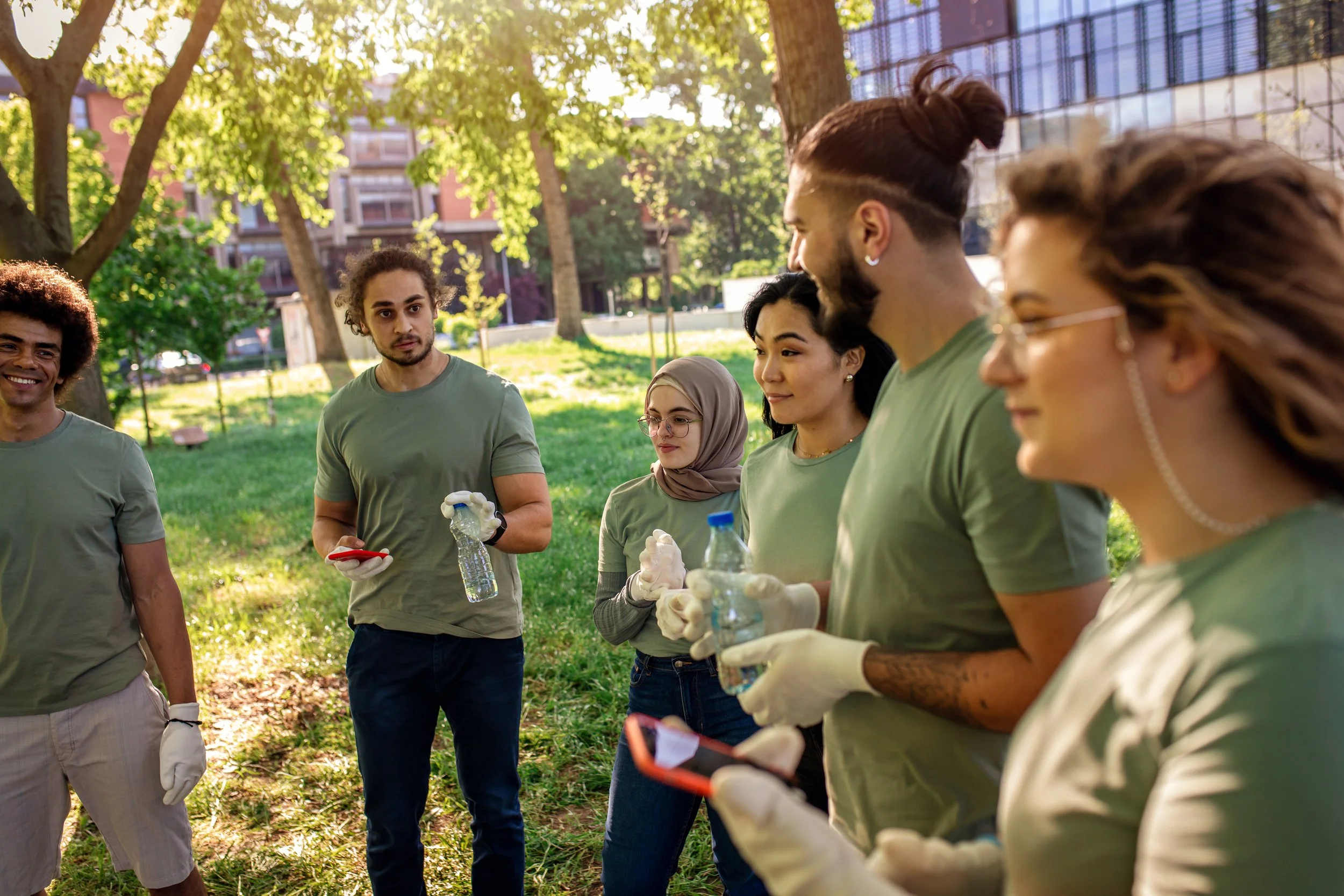 Group of diverse volunteers in green shirts gathering in a park for a community cleanup, holding water bottles and gloves, with trees and buildings in the background.