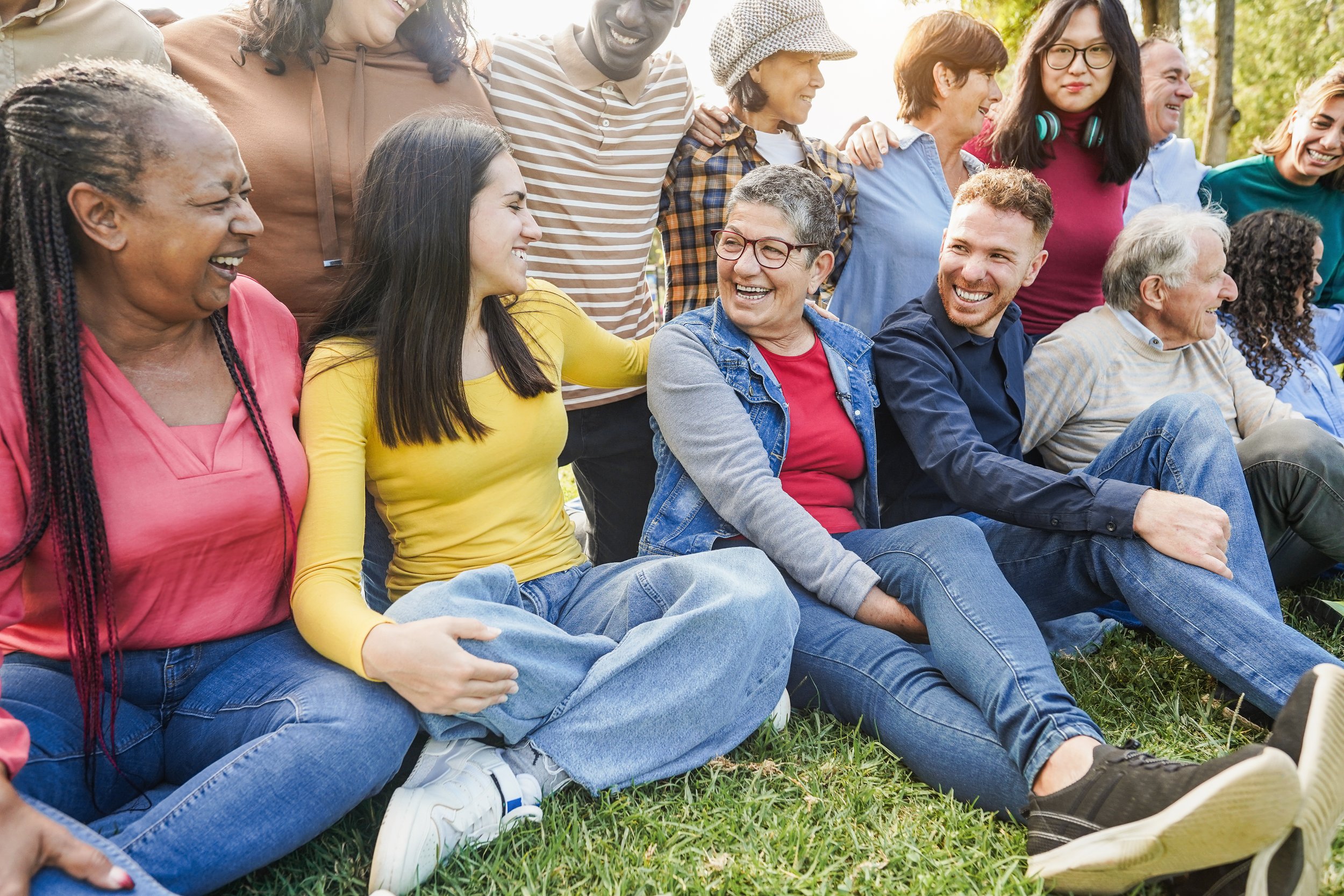 A diverse group of people sitting and standing outdoors, smiling and laughing, enjoying a sunny day in a park.