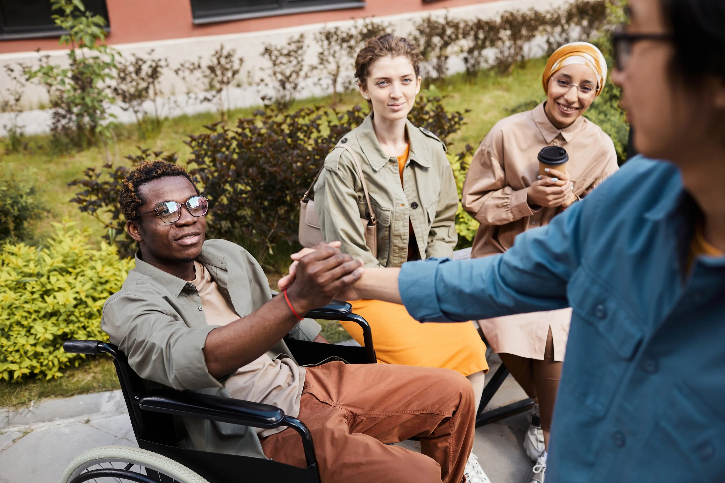 Four diverse young adults outdoors, one in a wheelchair shaking hands with another, while the others observe and smile, in a park setting with greenery and trees.