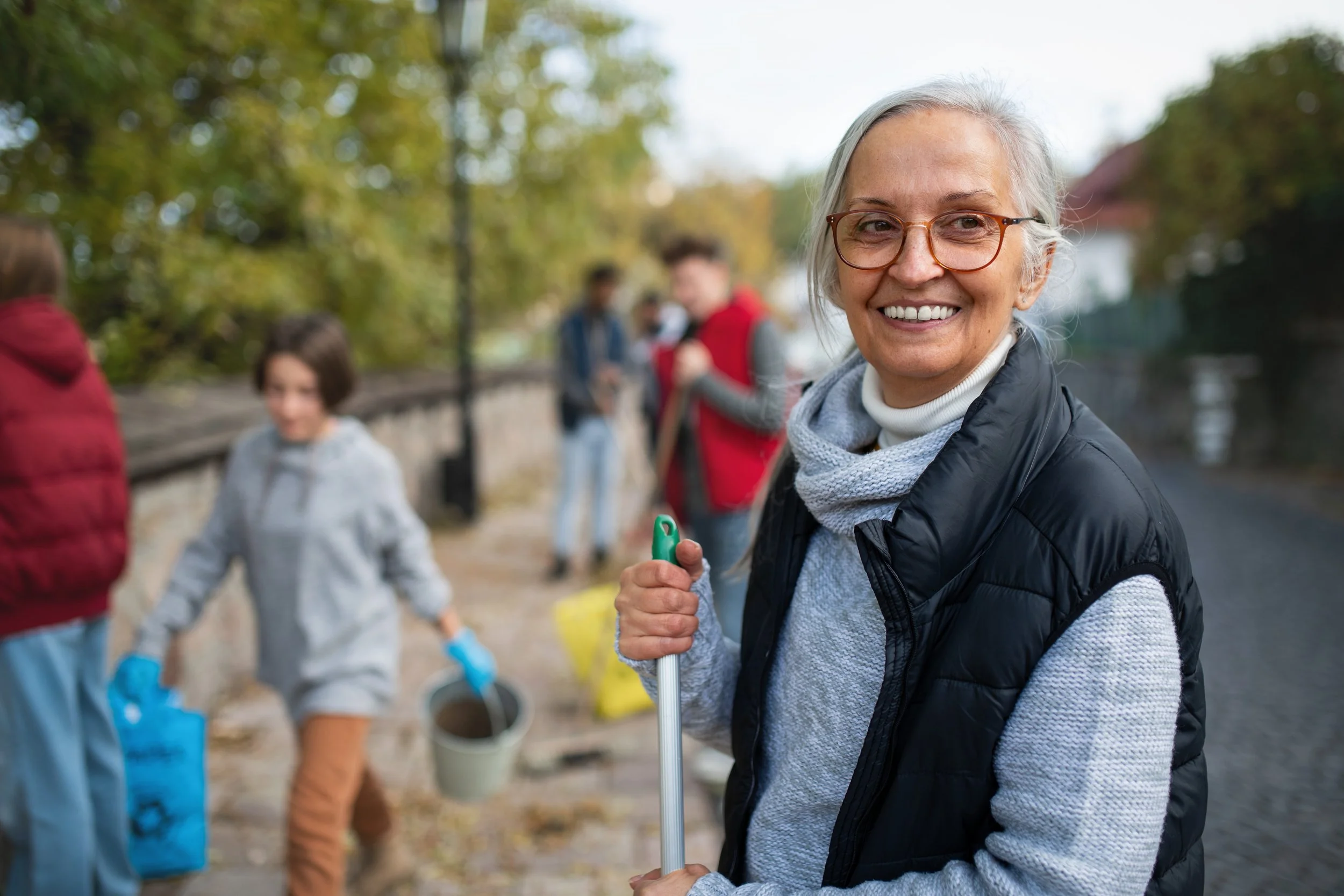 Smiling elderly woman with glasses holding a trash picker on a park pathway with a group of volunteers cleaning up in the background.