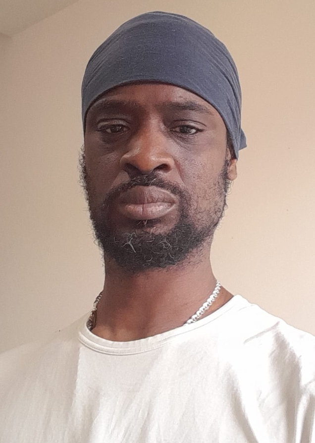 A man with dark skin wearing a gray head covering, a white shirt, and a silver chain necklace, standing against a plain beige wall.
