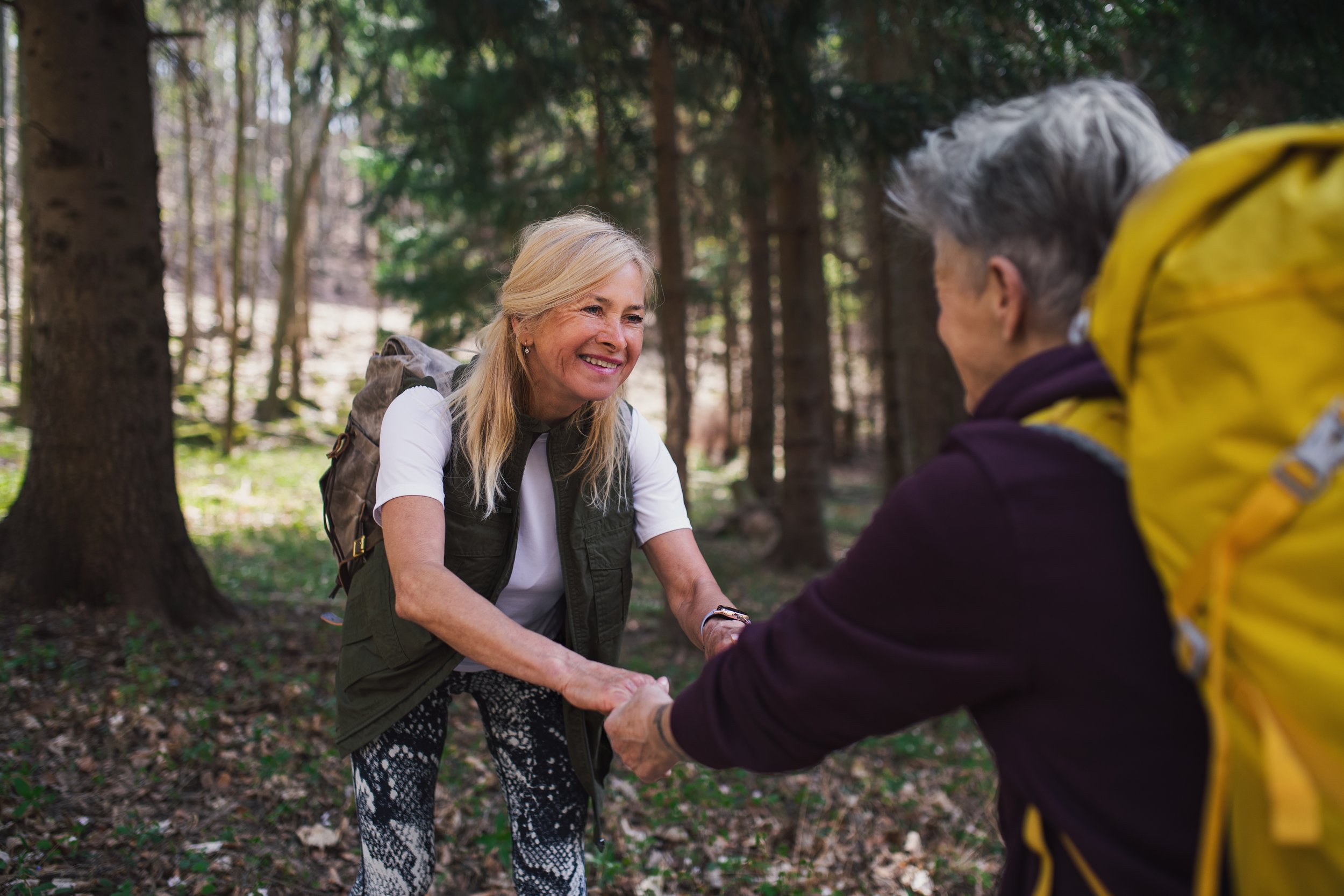 A woman with blonde hair smiling and holding hands with an older woman with gray hair in a forest.