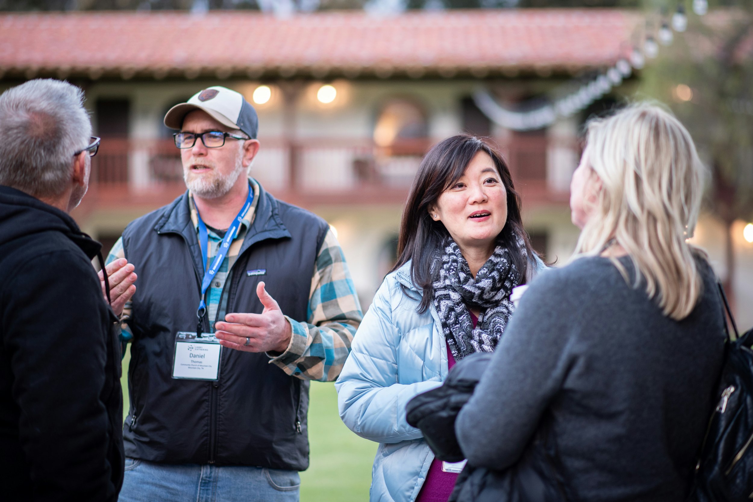 Four people engaged in conversation outdoors during evening, with a house and string lights in background.