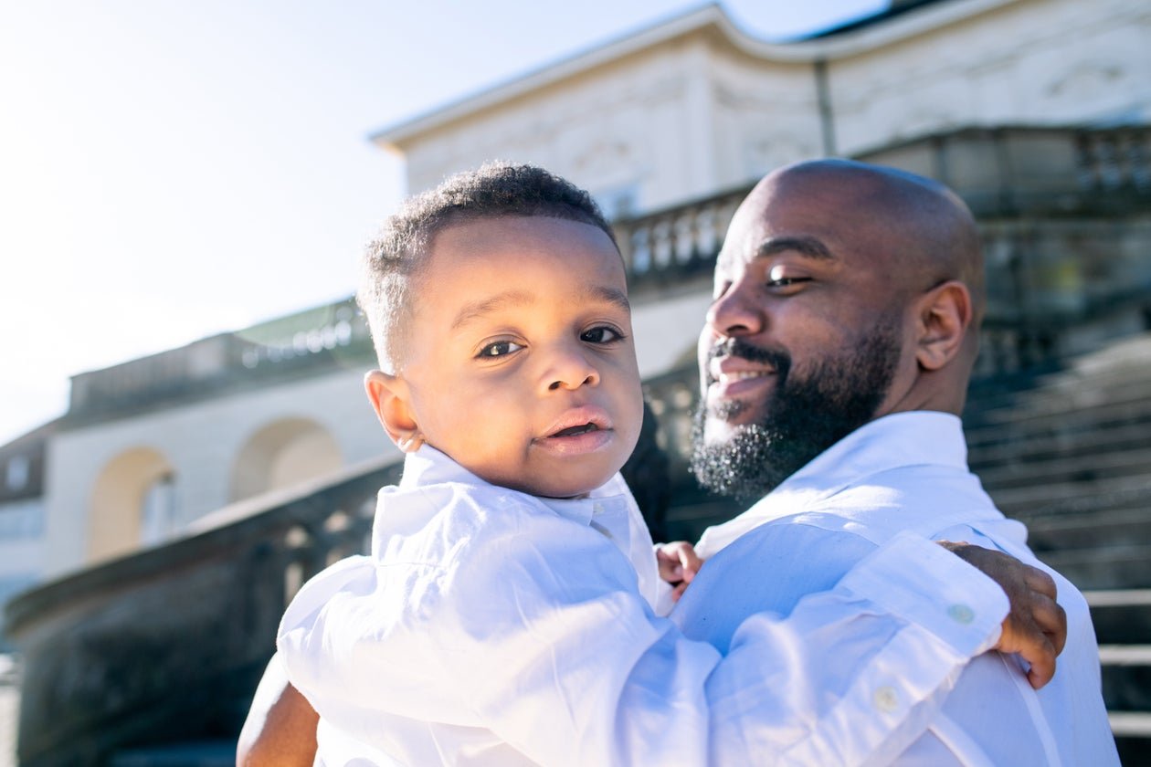 A man holding a young boy outside a white building with stairs, both wearing white shirts, smiling, with the sun shining.