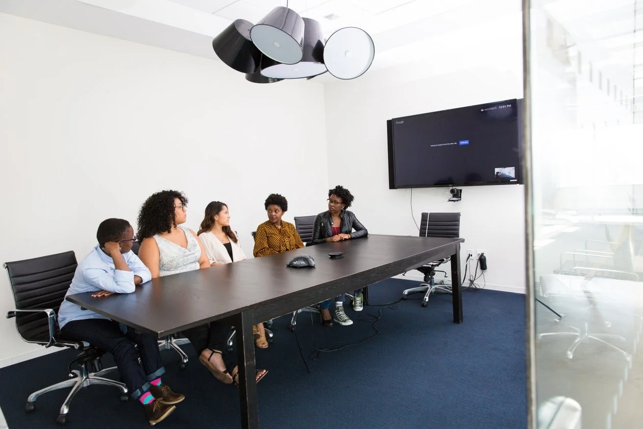 Five women sitting at a conference table in a modern meeting room, with a large screen TV on the wall and a conference phone on the table.