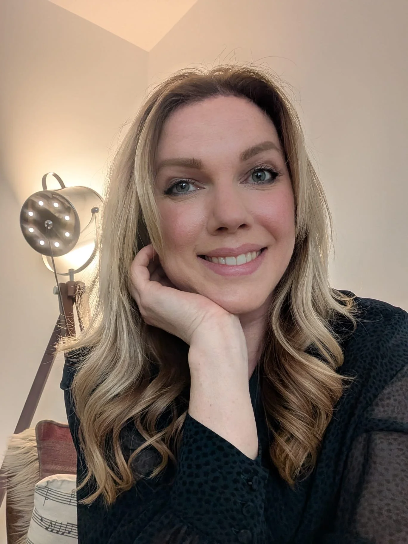 A woman with long wavy blonde hair and blue eyes smiling, resting her hand on her chin, seated in a cozy room with warm lighting, Wildflower Neuro Scarborough North Yorkshire