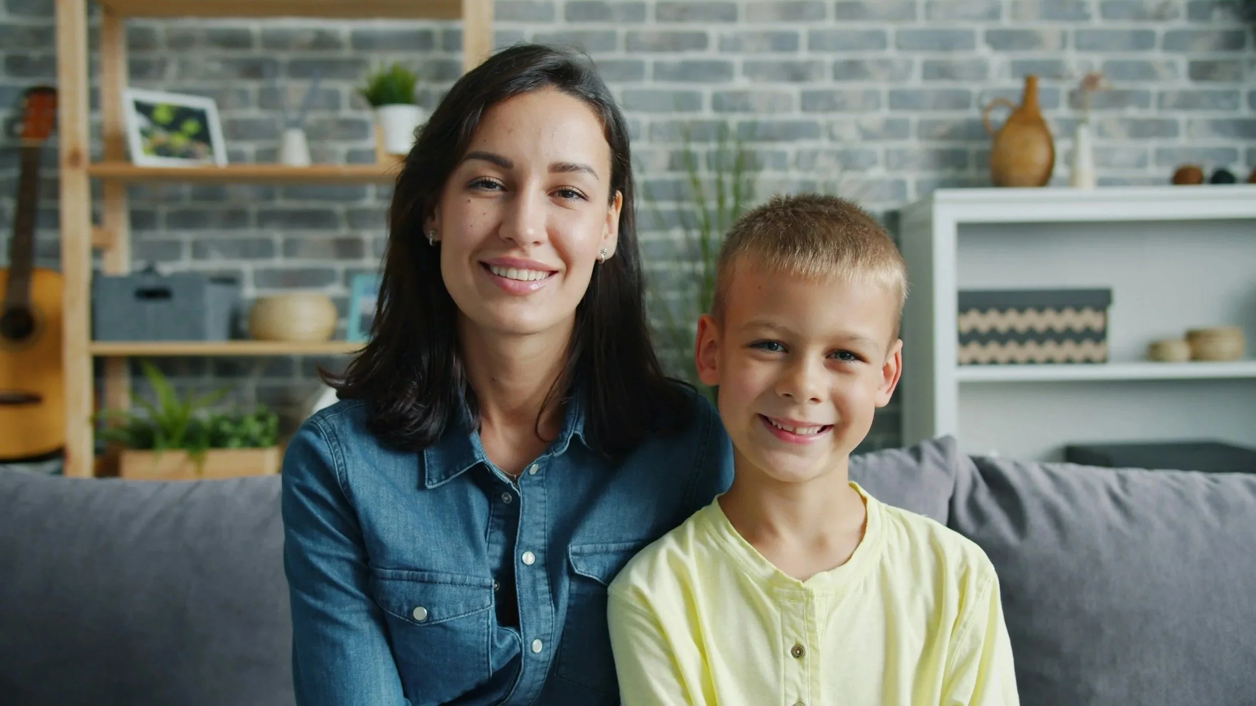A woman with dark hair and a boy with blonde hair smiling together inside a home with a brick wall, decorated with plants, shelves, and vases in the background. Wildflower Neuro Scarborough North Yorkshire