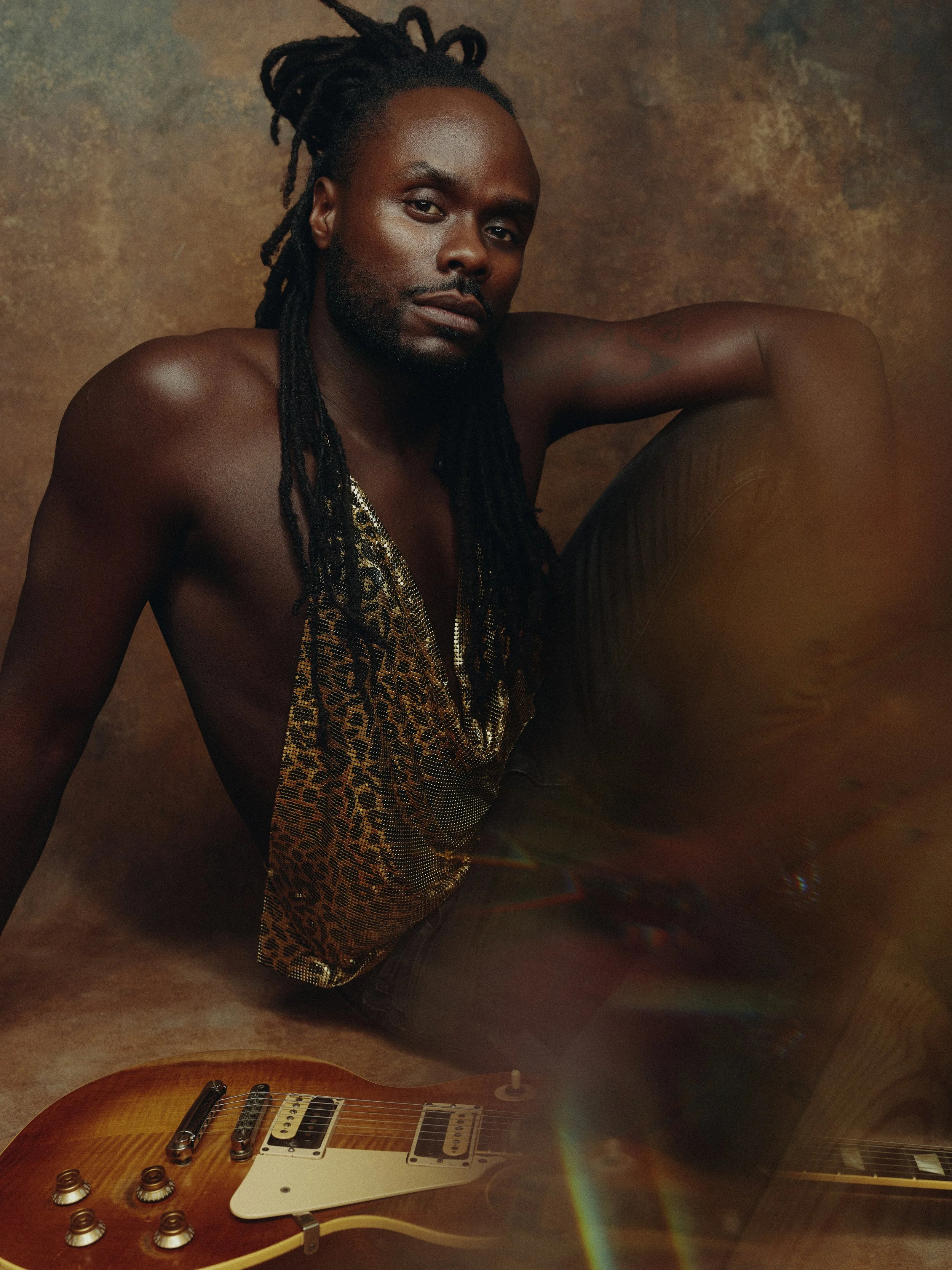 A shirtless man with dreadlocks sitting on the floor, leaning on one arm, with an electric guitar nearby, against a textured brown background.