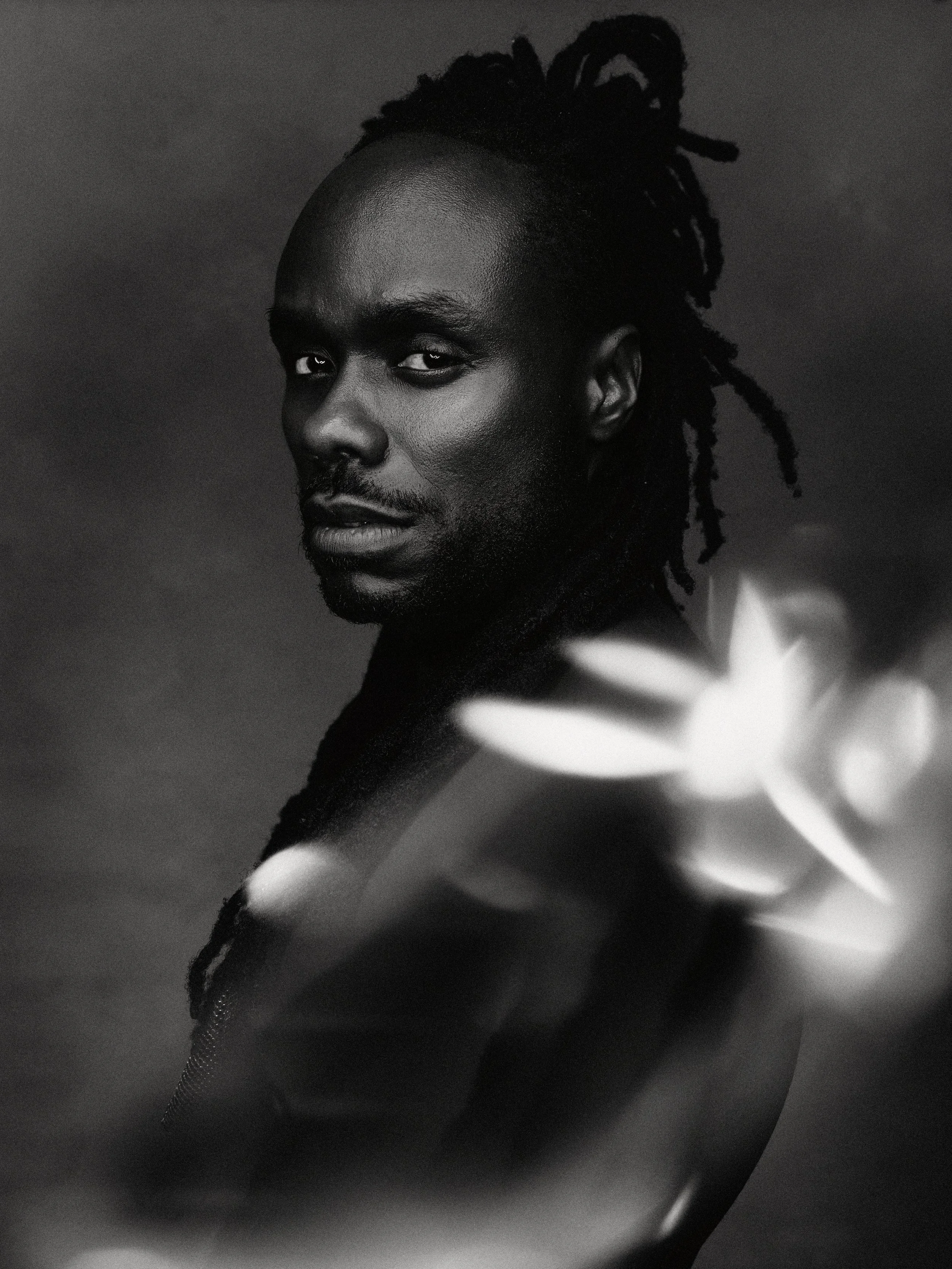 Black and white portrait of a man with dreadlocks reaching past his shoulders, looking directly at the camera, with a blurred light or object in the foreground.