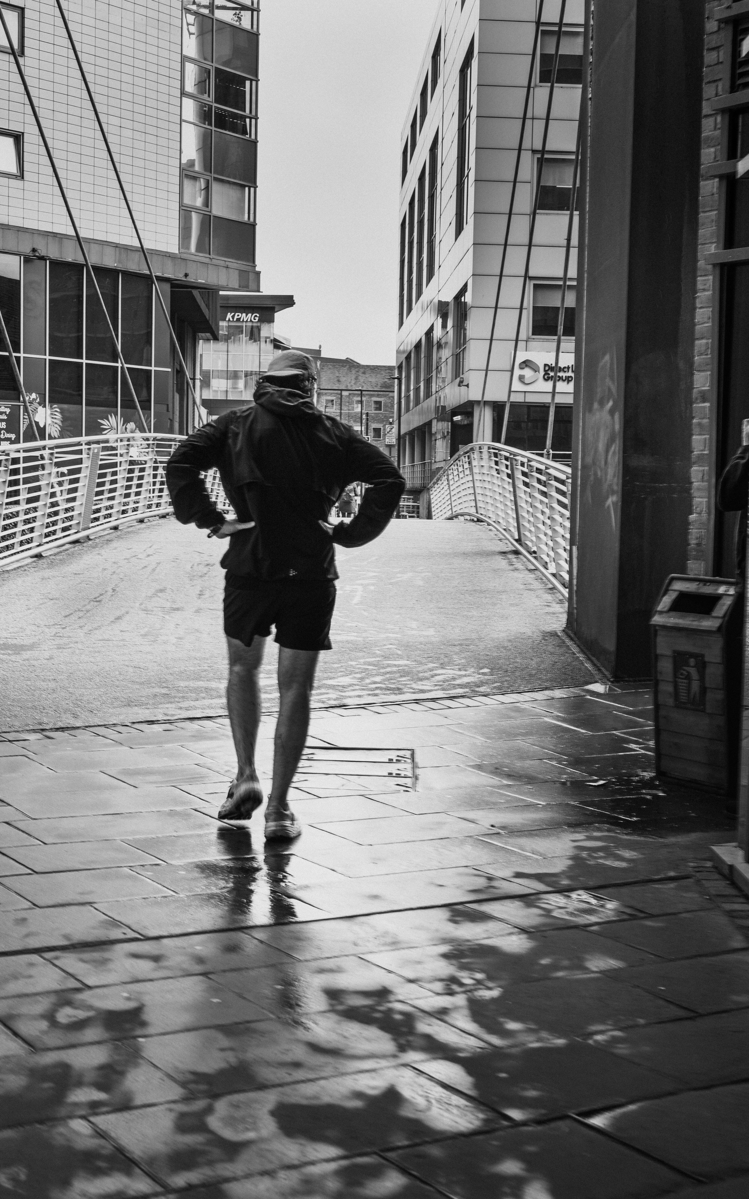 A person running in a city street, wearing a jacket and shorts, with reflections on wet pavement, surrounded by modern glass buildings.