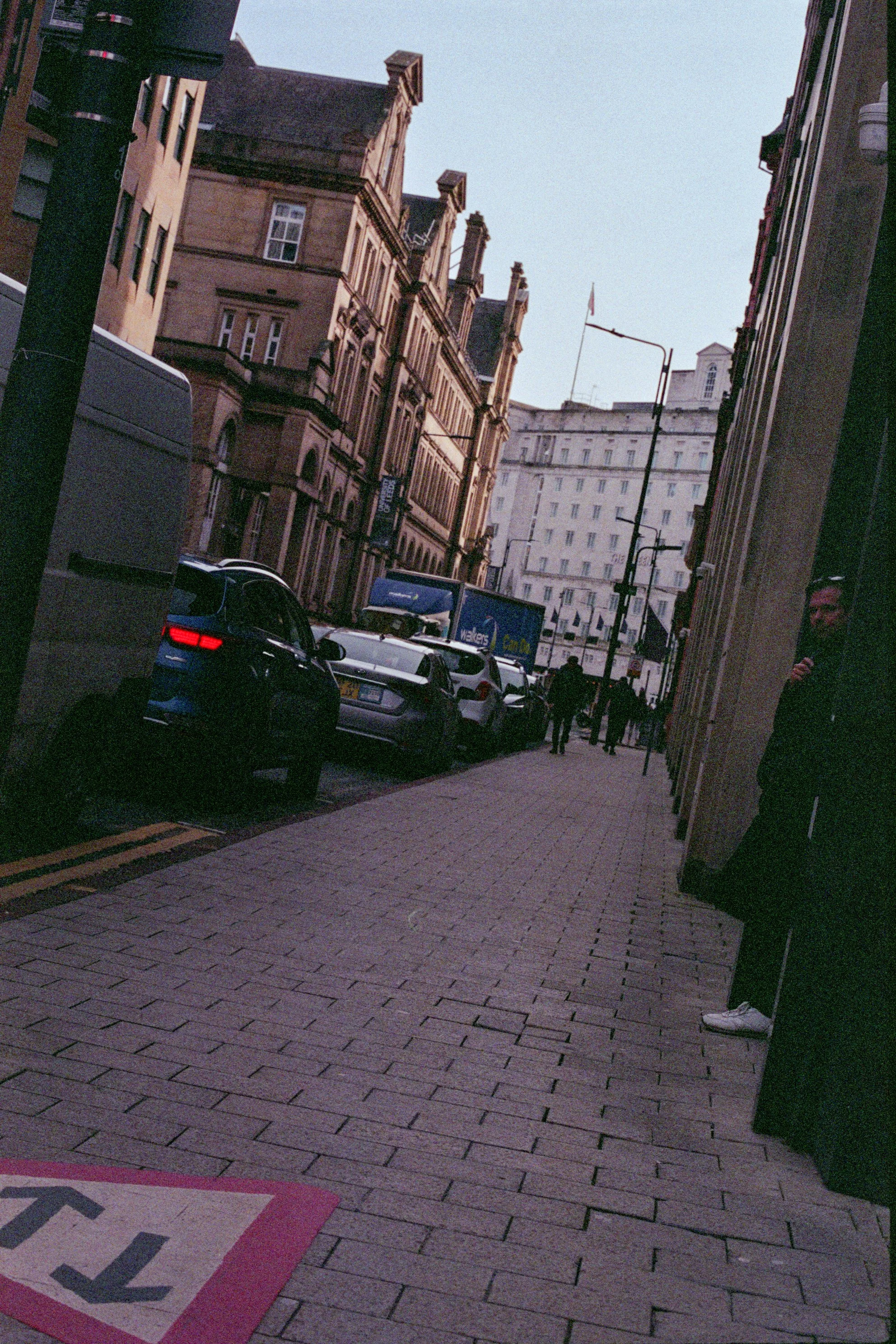 City street scene during dusk with parked cars along the sidewalk and pedestrians walking. Tall historic buildings line the street, with an overcast sky above.