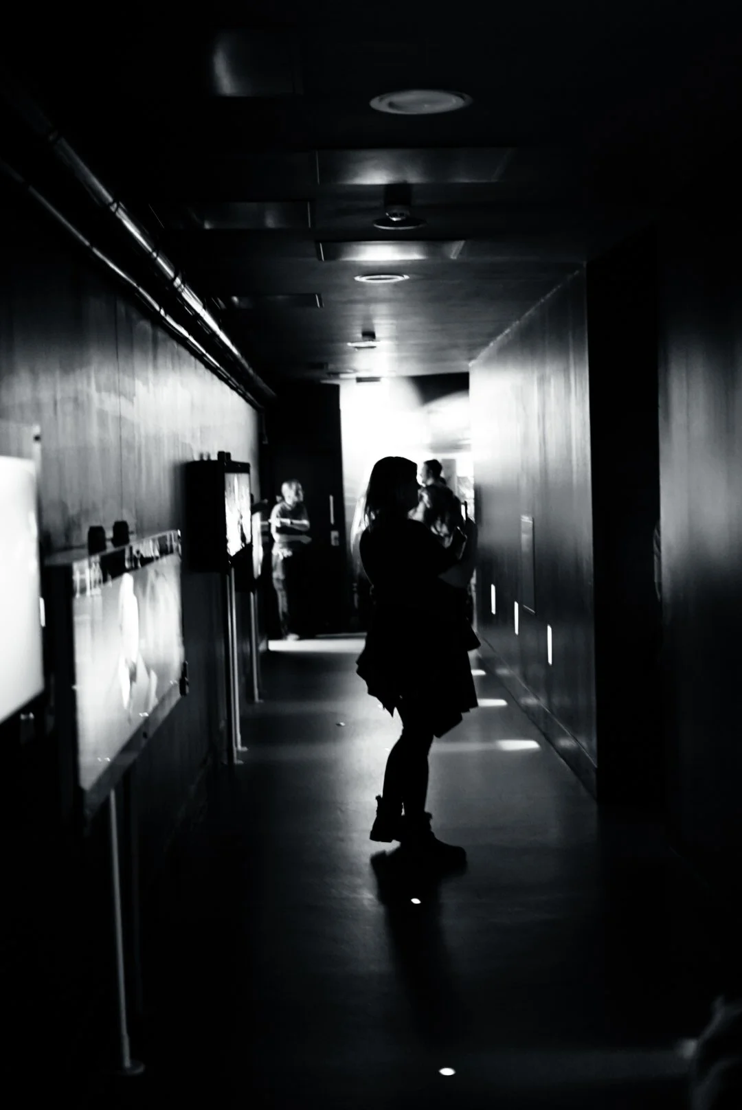 A black and white photo of a woman standing in a dark corridor with bright light behind her, while other people are seen in the background near the light.
