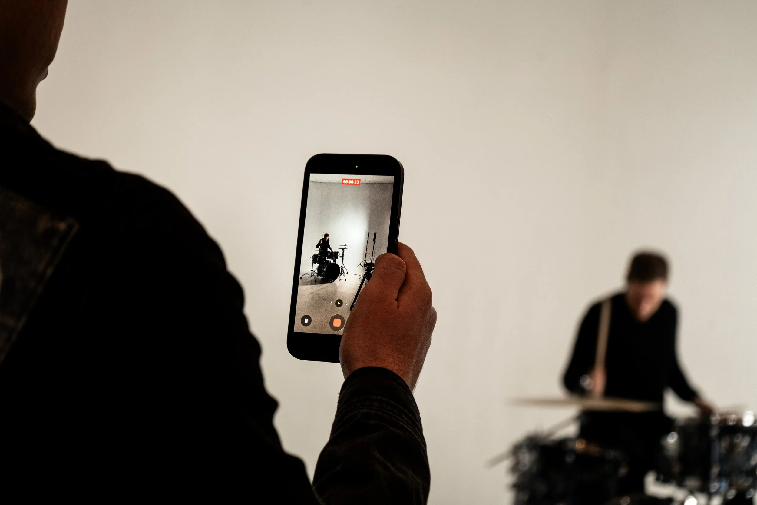 Person recording a musician playing drums in front of a plain white background on their smartphone.