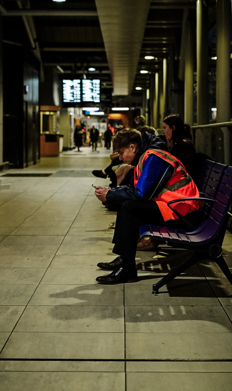 A young man in a red and blue jacket sitting on a bench in an airport terminal, looking at his phone, with other people sitting nearby.