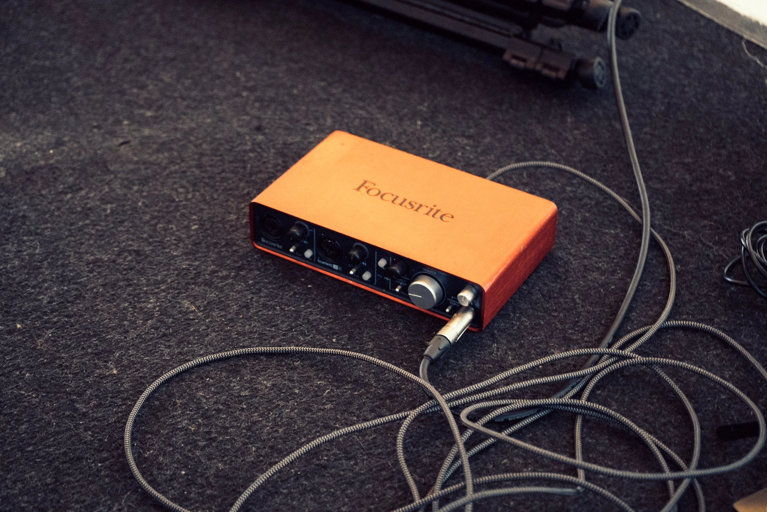 orange Focusrite audio interface with connected cables on a black carpeted floor.