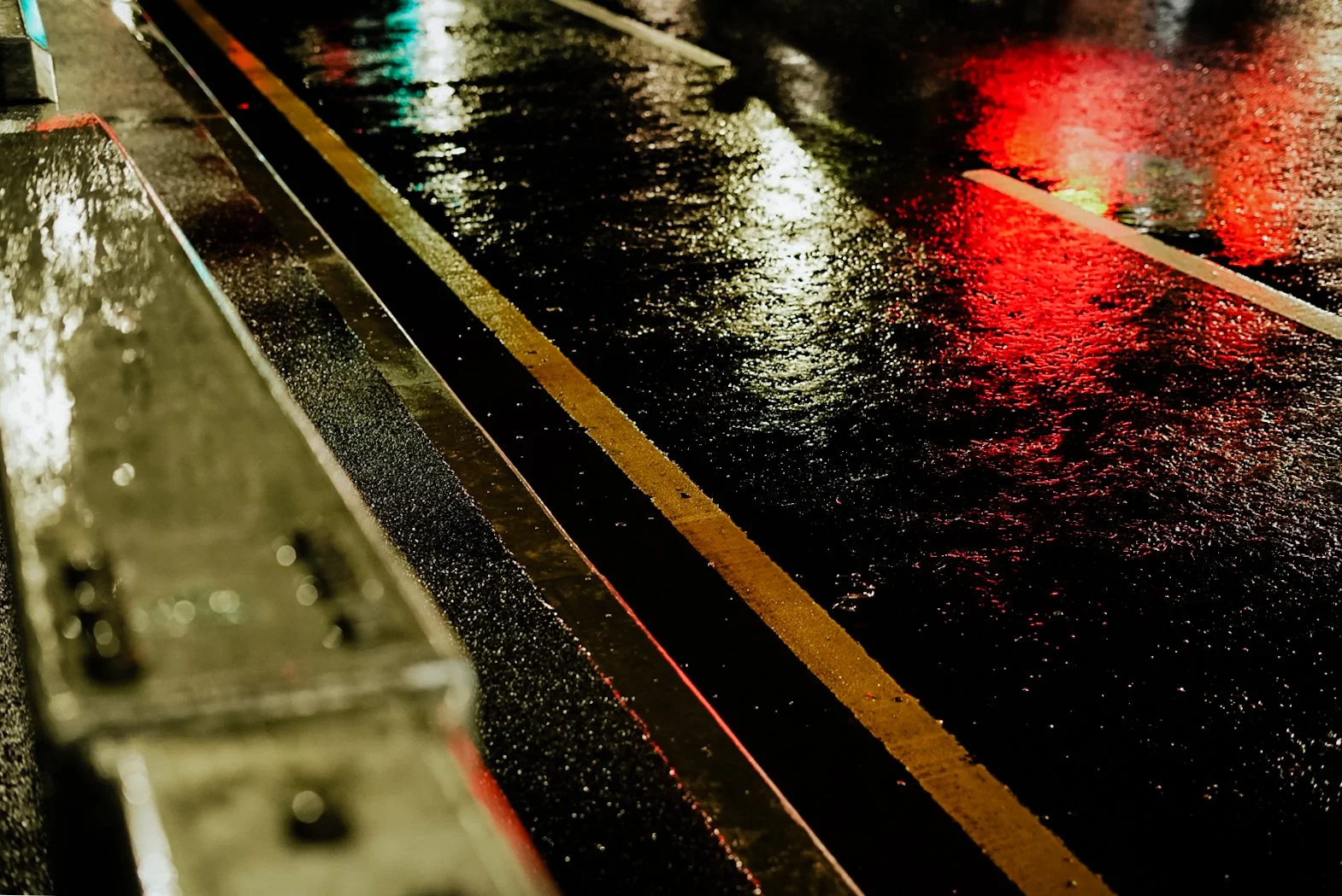 Wet street with reflections of red and white lights, with a metal barrier and road markings visible.