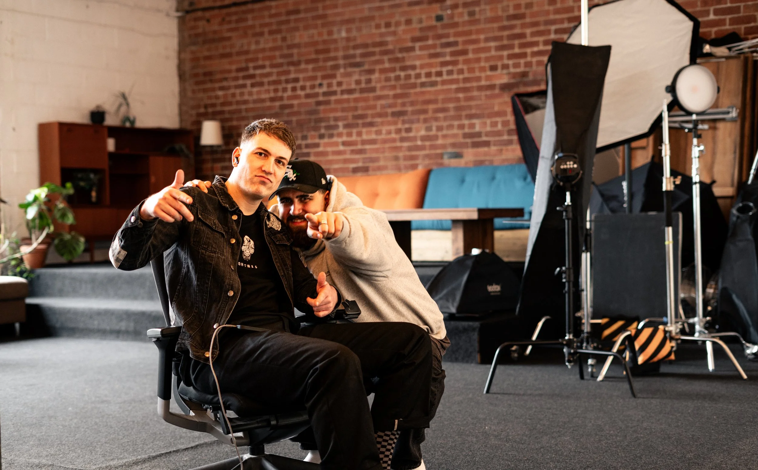 Two men in a photography studio, one sitting on a chair and the other leaning in, pointing at the camera. Studio equipment and a brick wall are visible in the background.