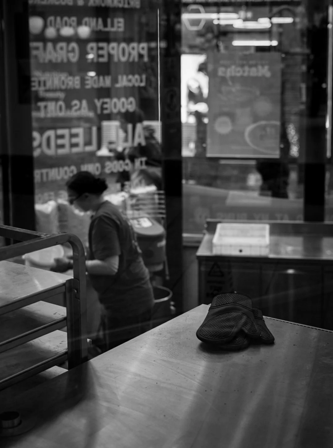 Black and white photo showing a kitchen or pizza place with a dough scraper or glove on a table in the foreground. Through a glass window, a person wearing glasses and a T-shirt is seen preparing food, with several signs and reflections visible on th