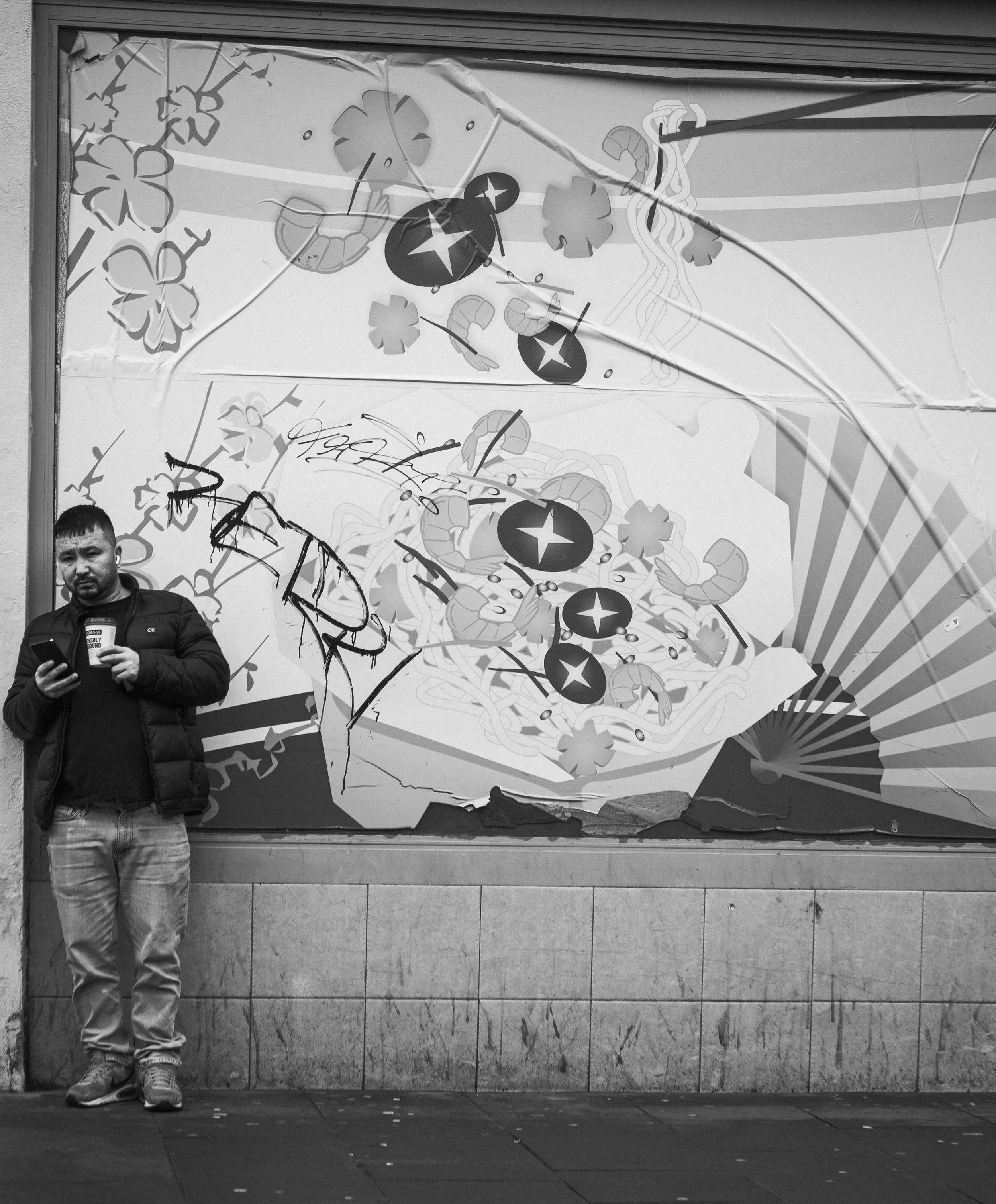 A man standing on a sidewalk in front of a store window mural showing spilled Asian noodles, with some graffiti on the glass.