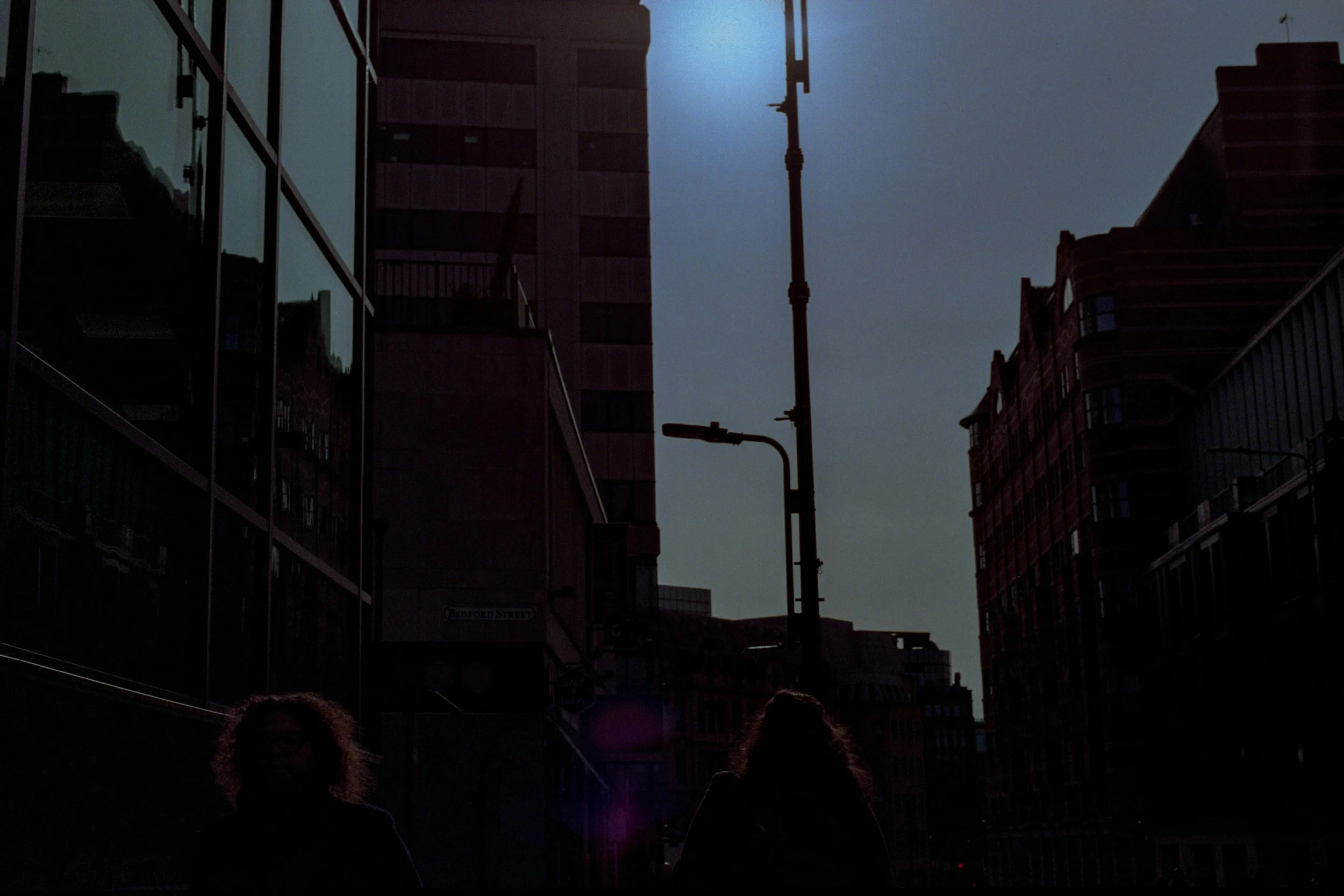 City street scene with tall buildings, a streetlight, and a cloudy sky. Silhouettes of two people walking.