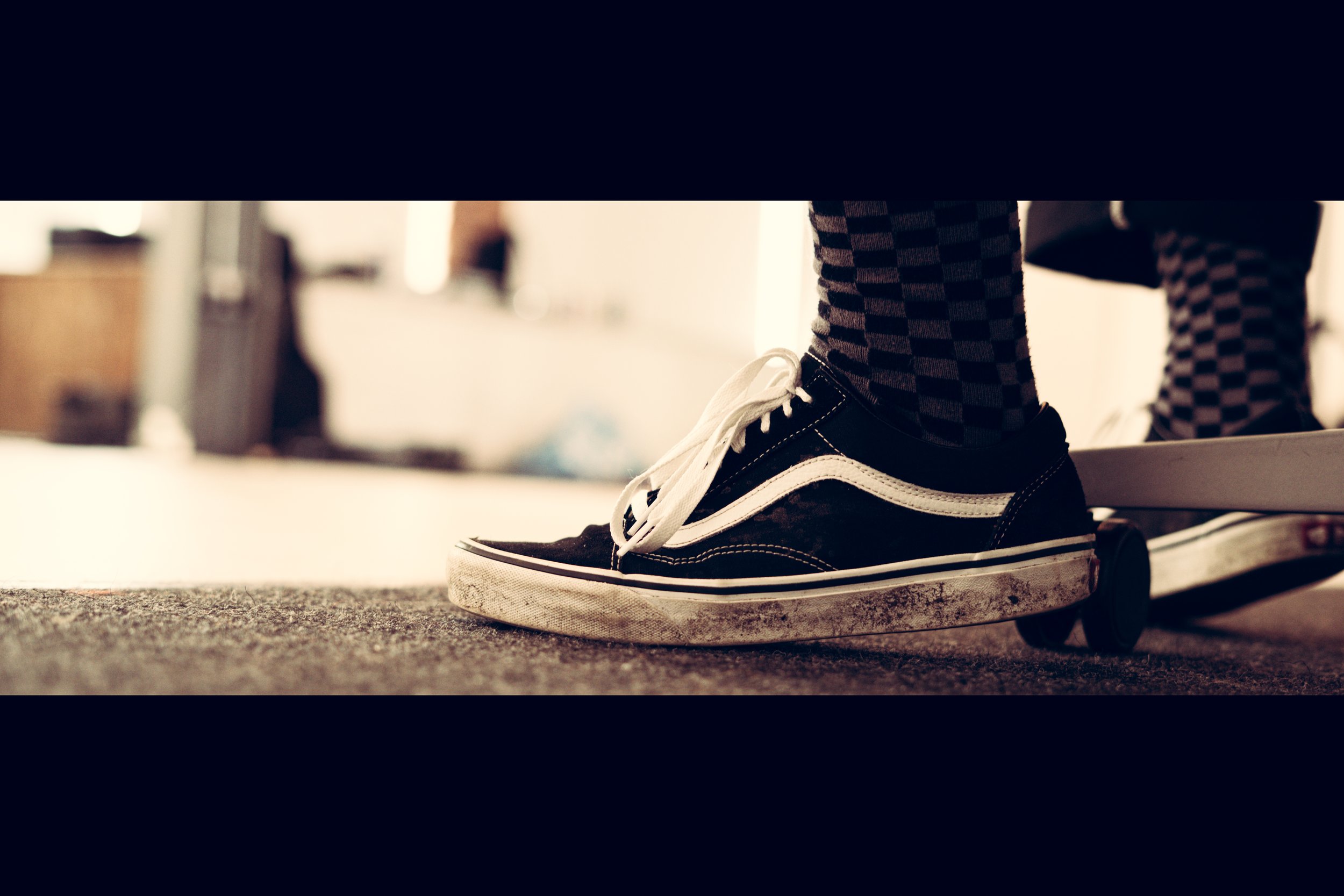 Close-up of a person with checkered socks and black Vans sneakers riding a skateboard on a carpeted floor.