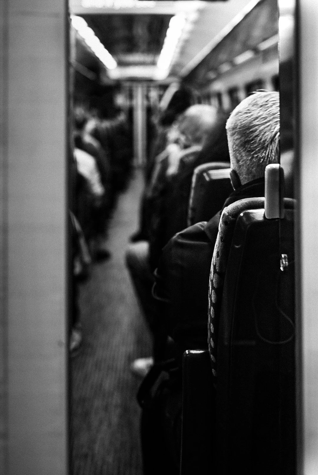 Black and white photo of the inside of a train or bus, showing passengers seated, facing forward, with one person with short, light-colored hair in the foreground.