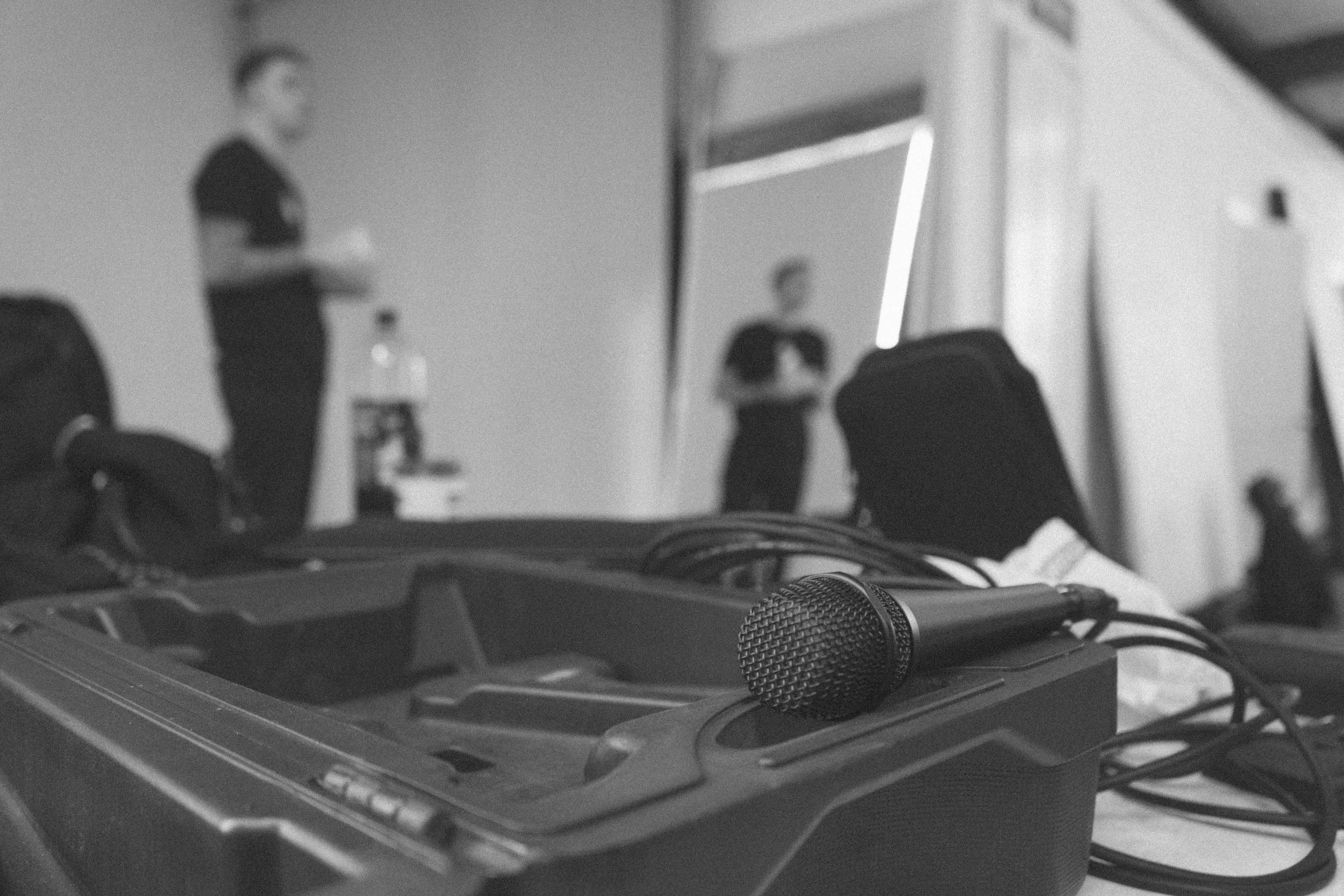 Close-up of a microphone on a table, with people and a screen visible in the background, likely at a conference or presentation.