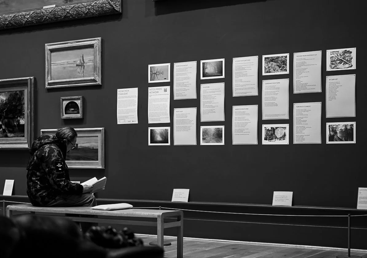 Person sitting on bench reading a book in an art gallery with framed paintings and informational displays on the wall.